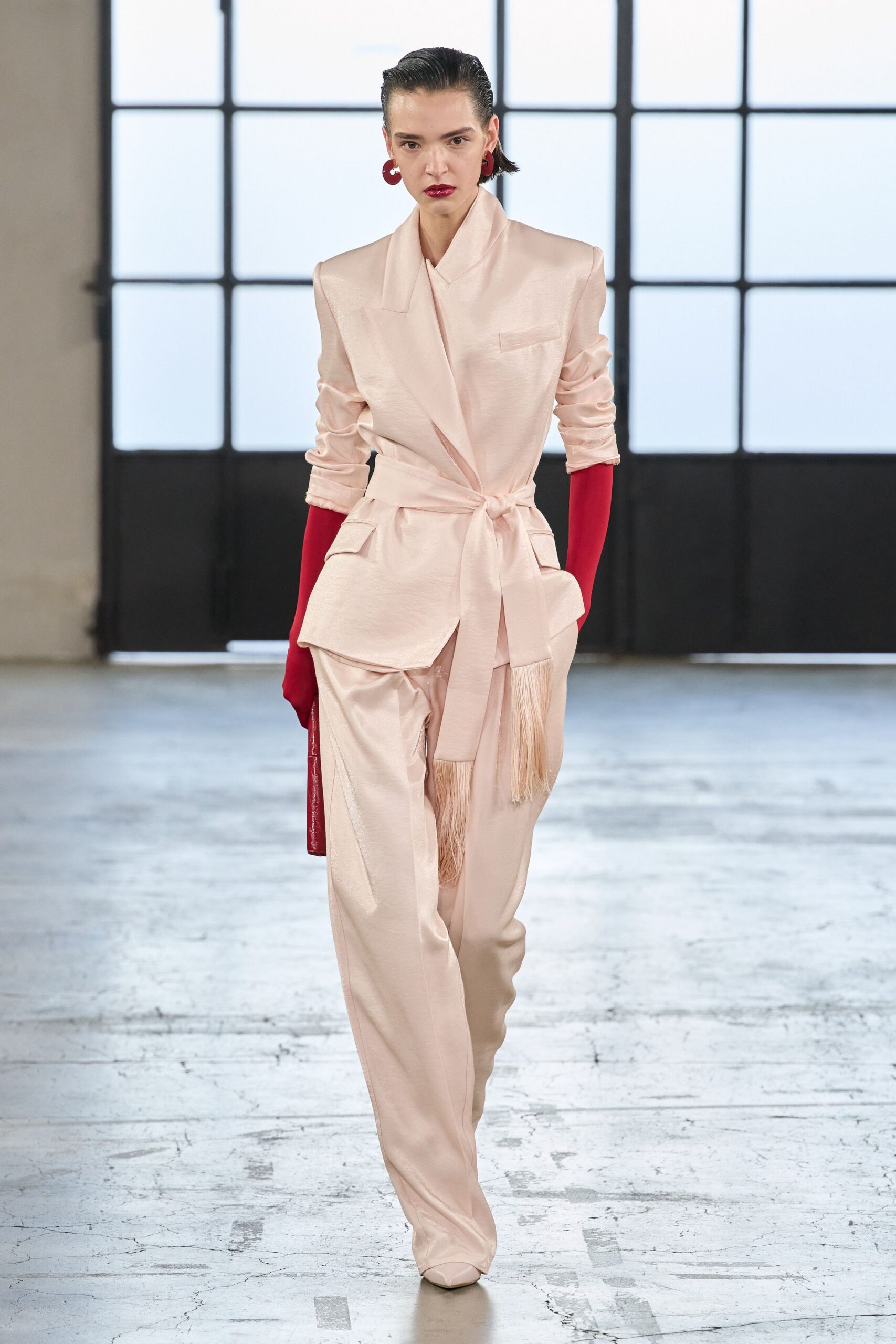 Large oxblood resin hoops. Blush satin blazer with padded shoulders and a fringed self-tie. Opera gloves in saturated crimson jersey. Loose blush satin trousers with a sharp front crease. Pointed boots disappearing under the hem. Walking into a wedding breakfast after spilling the first glass of red.