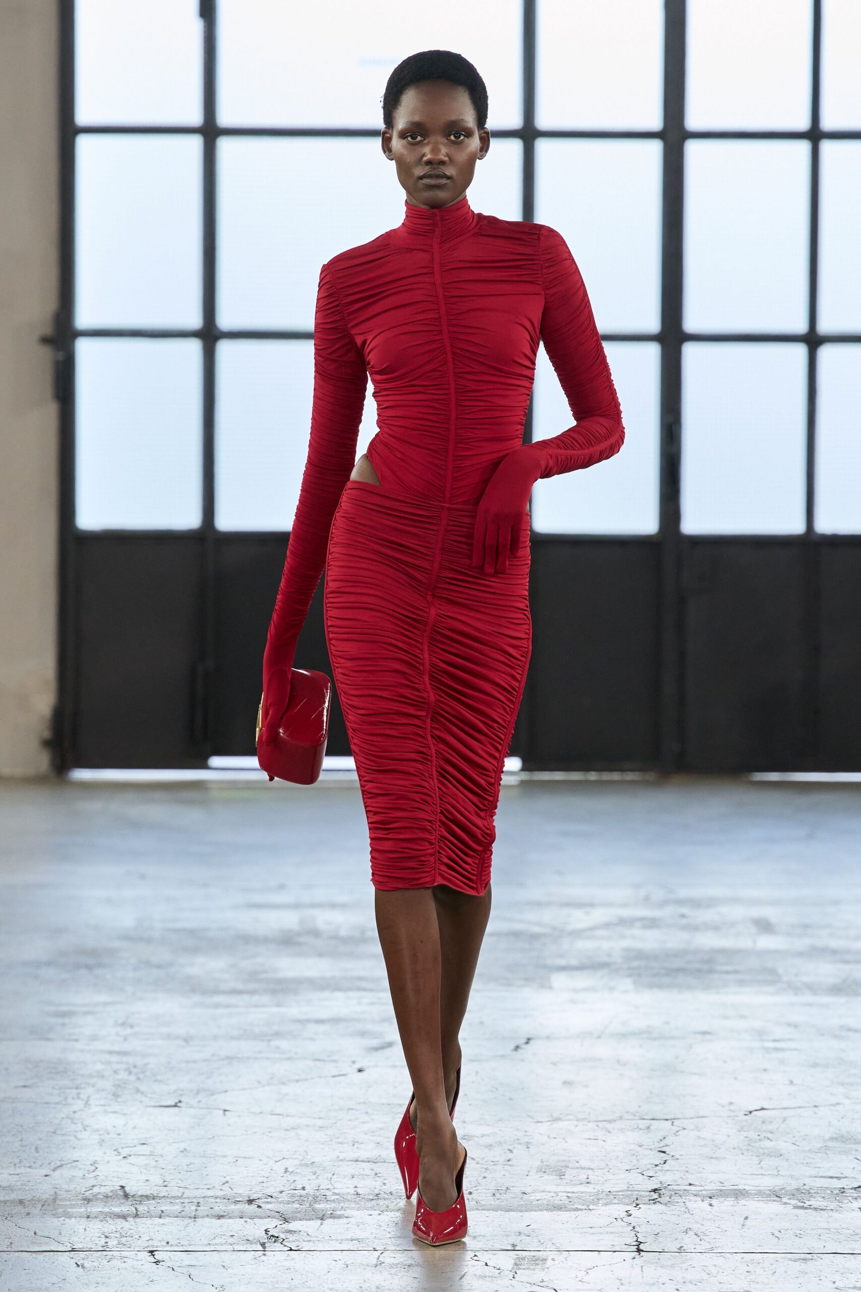 Ruched scarlet jersey dress with a stand collar and hip cutout. Built-in gloves extending from the ruched sleeves. Glossy red box clutch with a gold corner clasp. Red patent pumps, sharply pointed at the toe. Walking into a winter wedding reception after the church bells stopped.