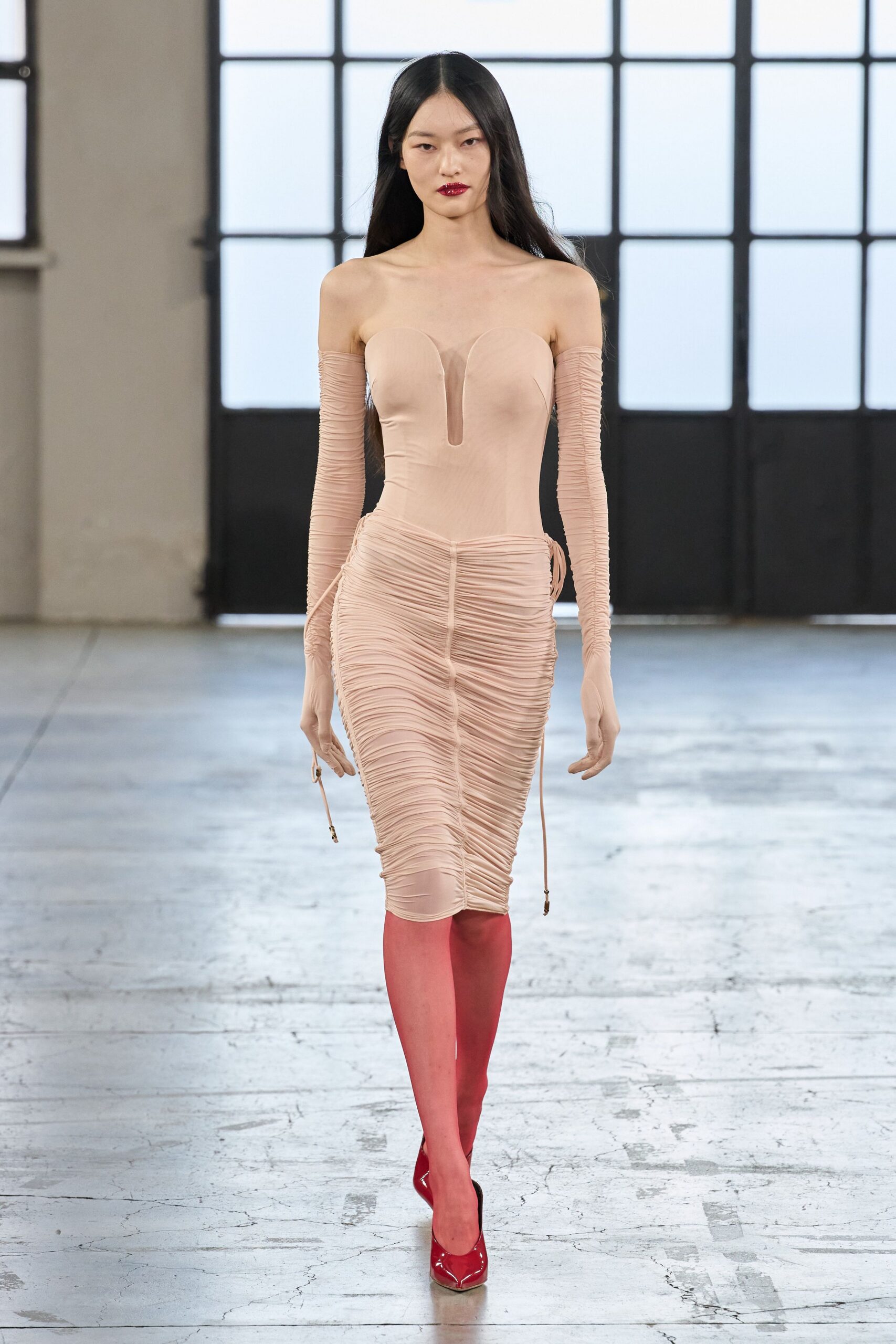 Blush stretch dress, plunged U notch and dense ruching to the knee. Detached ruched sleeves ending in dangling drawcord tips. Rose-pink sheer tights. Patent crimson pumps with squared throats. Walking across a cold loft floor after the radiator finally kicks in.