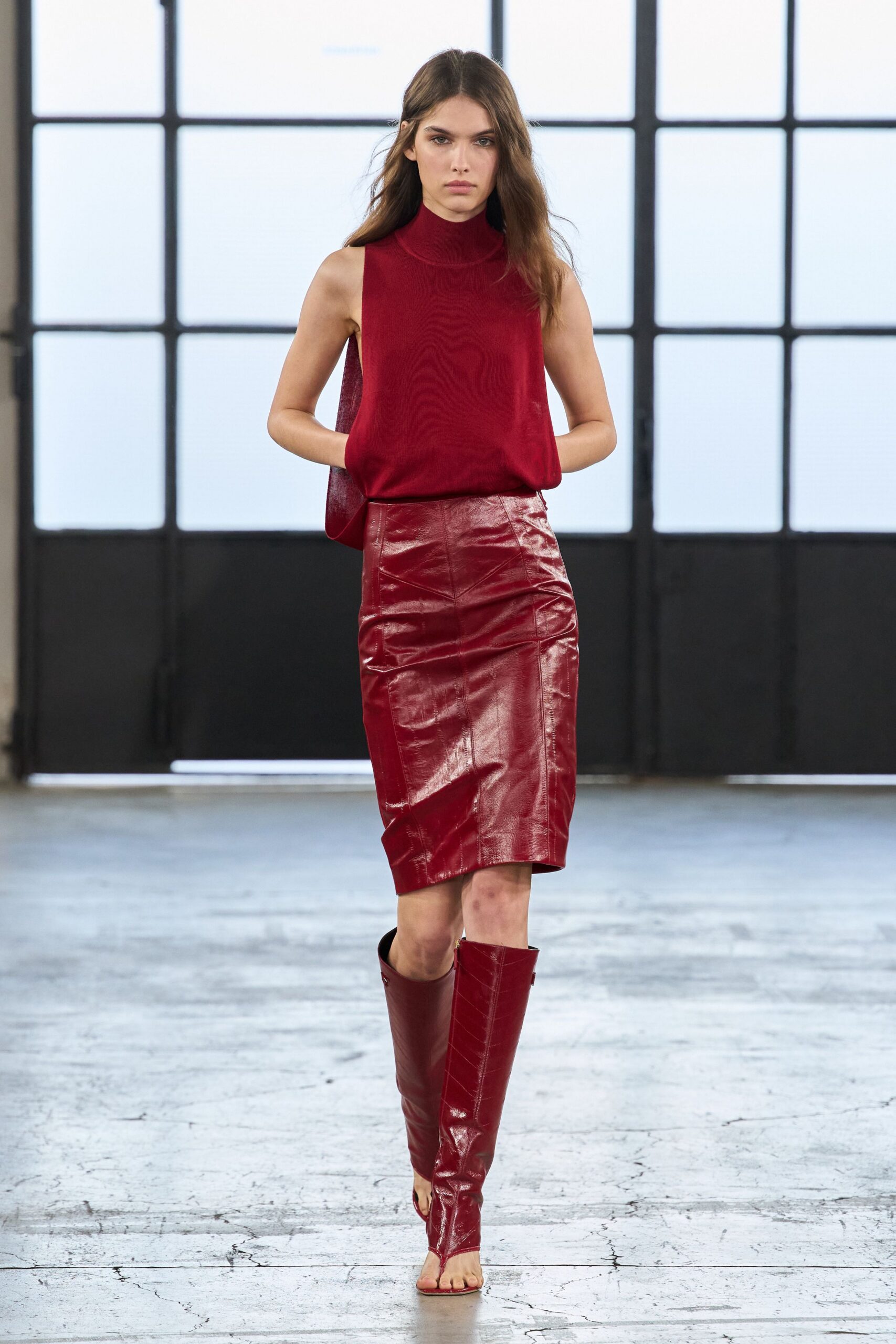 Dark cherry sleeveless knit with a rolled mock neck and dropped armholes. Glossy leather pencil skirt, paneled and wrinkled through the hip. Matching open-toe leather boots, one seam running straight to the ankle. Crossing a warehouse office at dusk, still carrying the heat outside.
