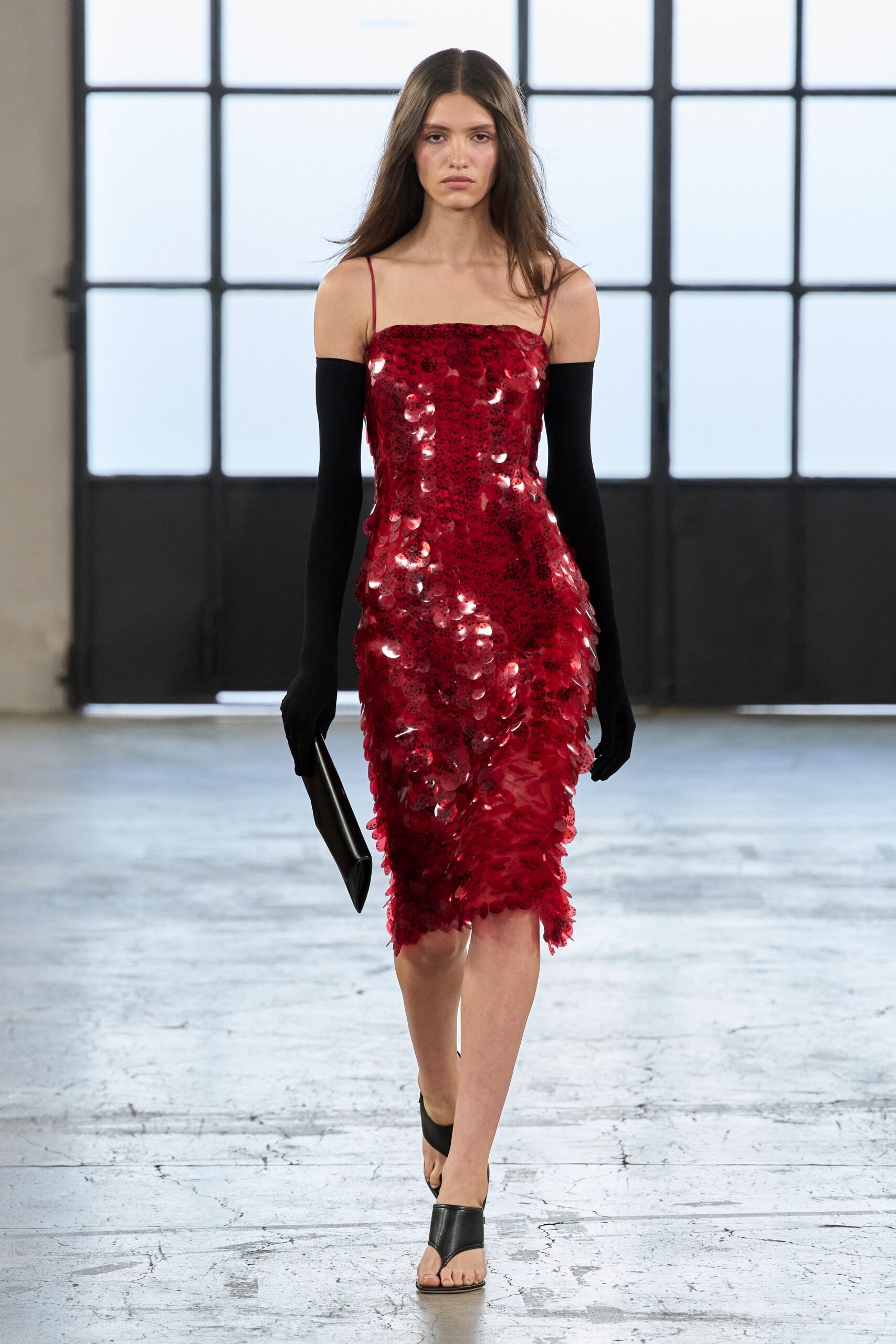 Crimson slip dress crusted in oversized paillettes, hem flickering with petal-like discs. Black opera gloves worn slouched off the shoulders. Flat black leather clutch tucked under one hand. Black leather toe-loop sandals with a sharp vamp. Crossing a hotel lobby after midnight with lipstick still on the glass.