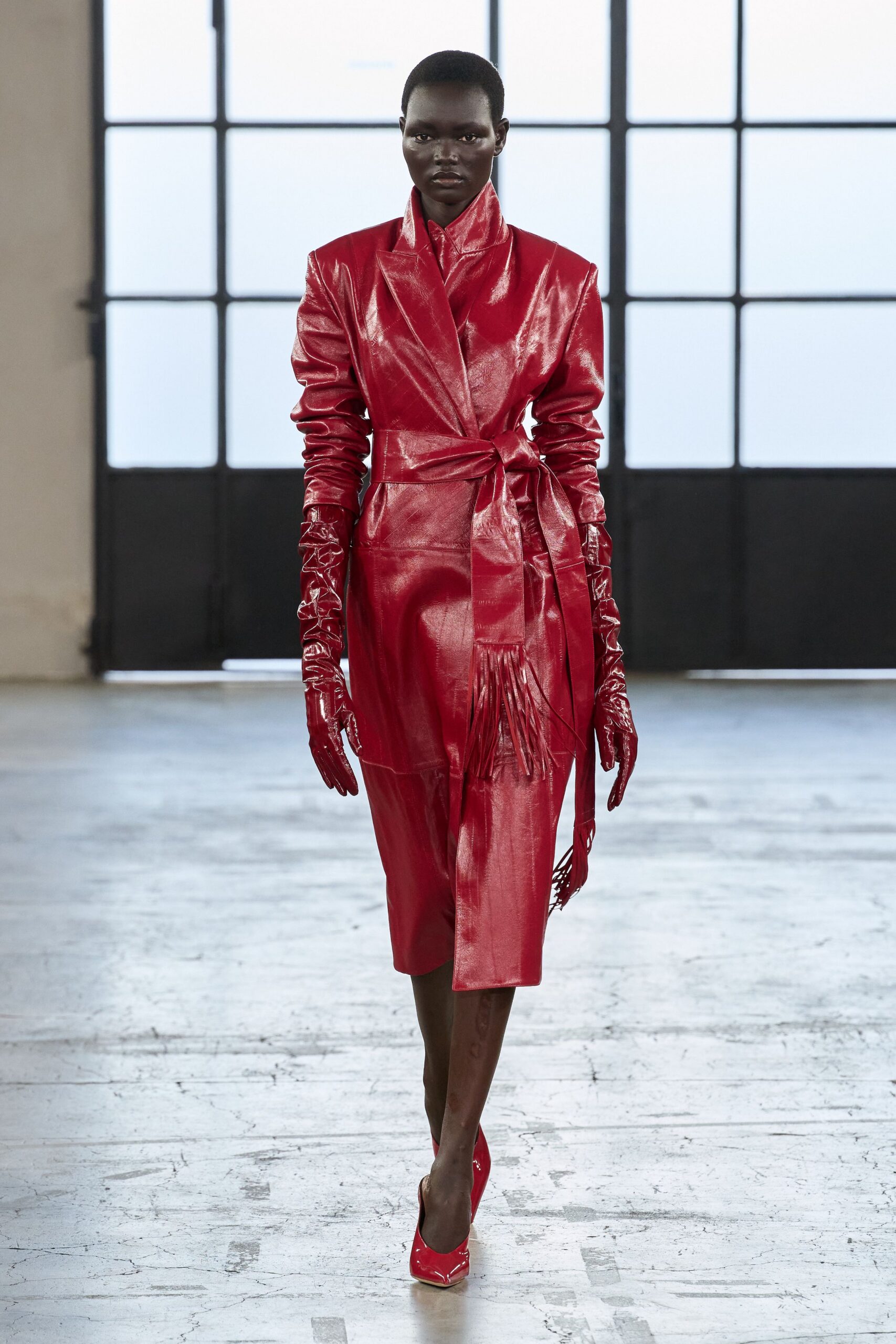 High-gloss scarlet leather coatdress with strong shoulders and a fringed wrap belt. Matching patent gloves, ruched in hard creases at the forearm. Pointed red patent pumps. For arriving first at a warehouse auction on a freezing morning.