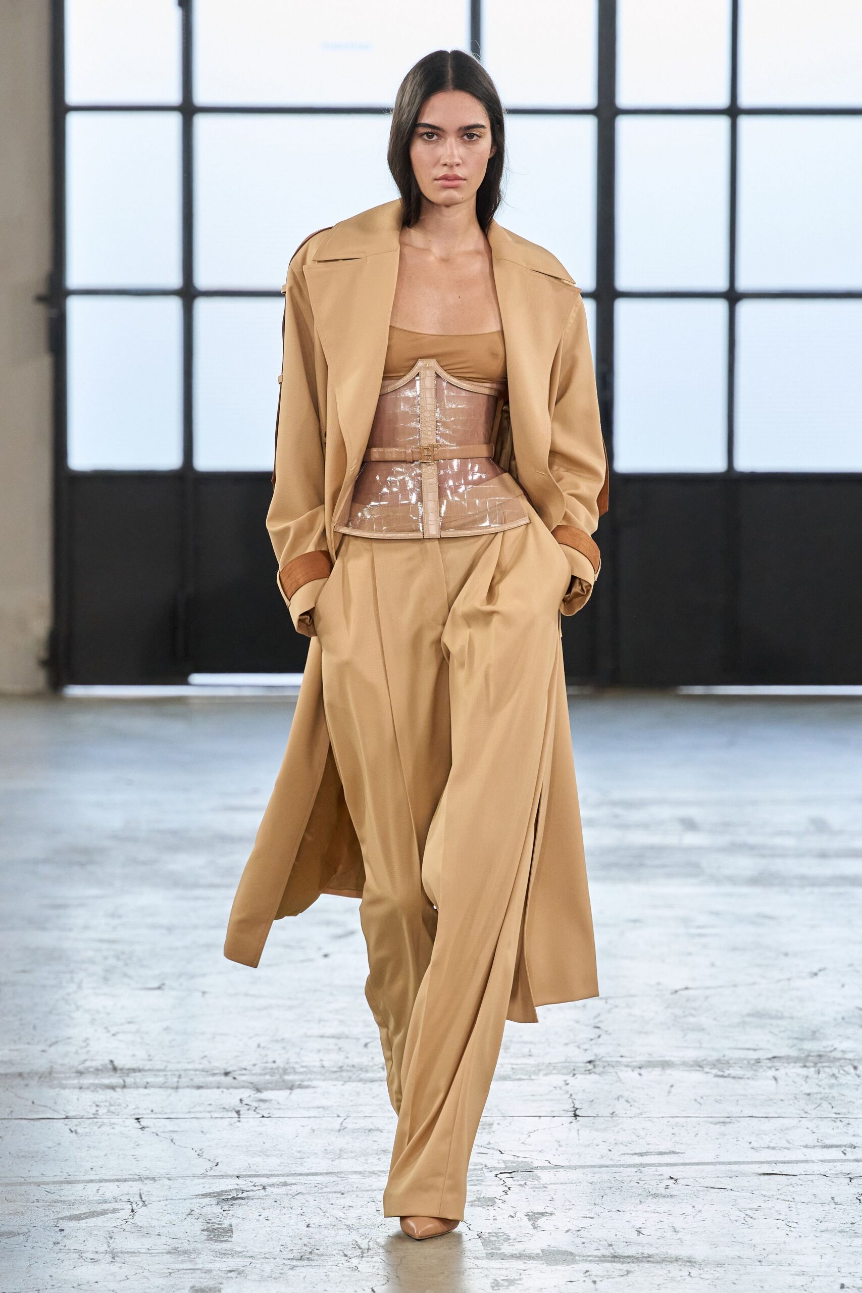 Camel duster coat with broad lapels and turned-back cinnamon cuffs. Chestnut bandeau cut straight across the bust. Clear vinyl corset with nude binding and a centered buckled strap. Camel trousers falling in long fluid pleats. Pointed camel pumps. For crossing a silent atrium before the office lights warm up.