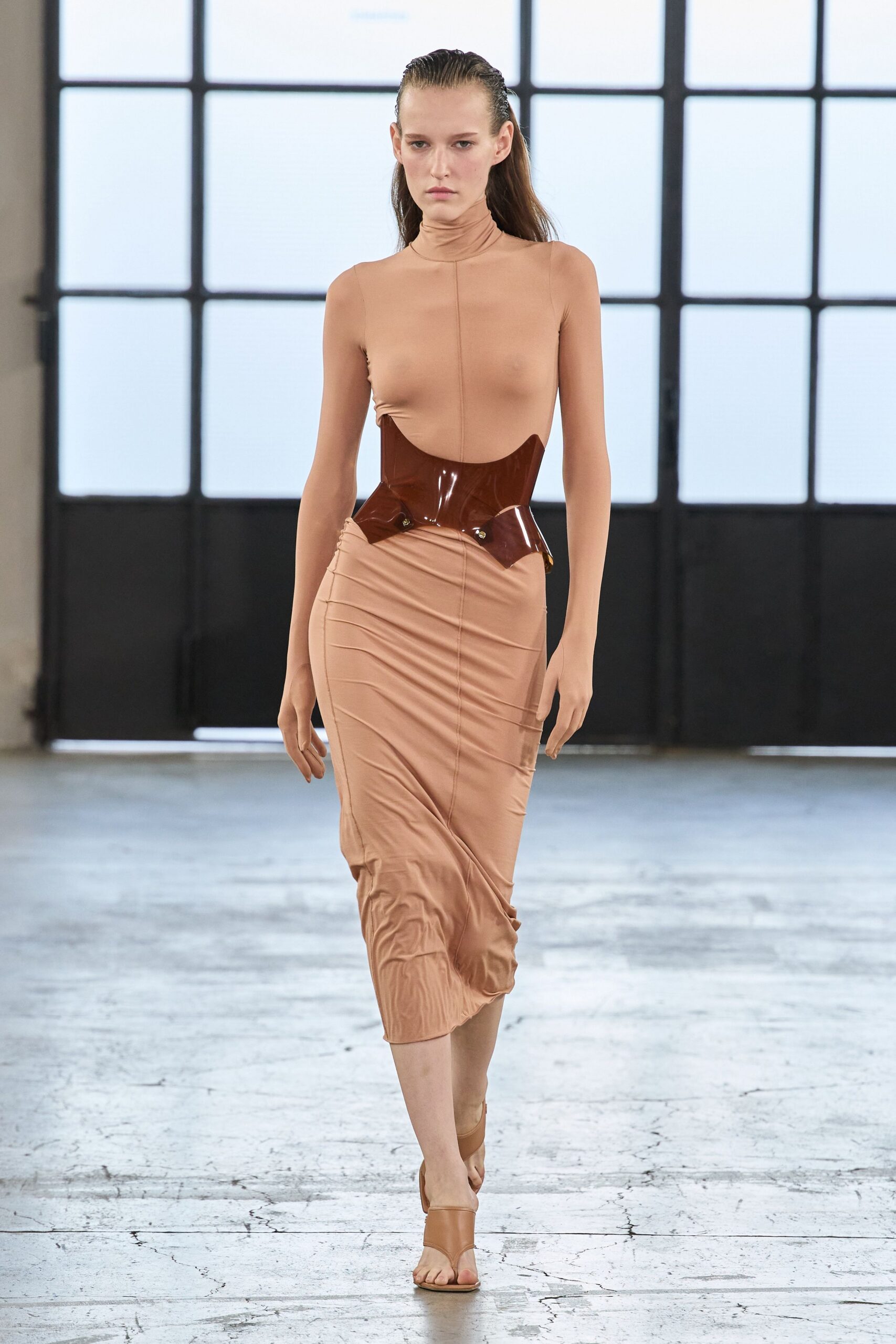 Nude stretch jersey dress with a rolled turtleneck and ruched midi skirt. Glossy oxblood molded corset belt, two small studs catching at the hip. Tan thong sandals with a narrow ankle strap. Crossing a concrete atrium while the rain still clings to the windows.