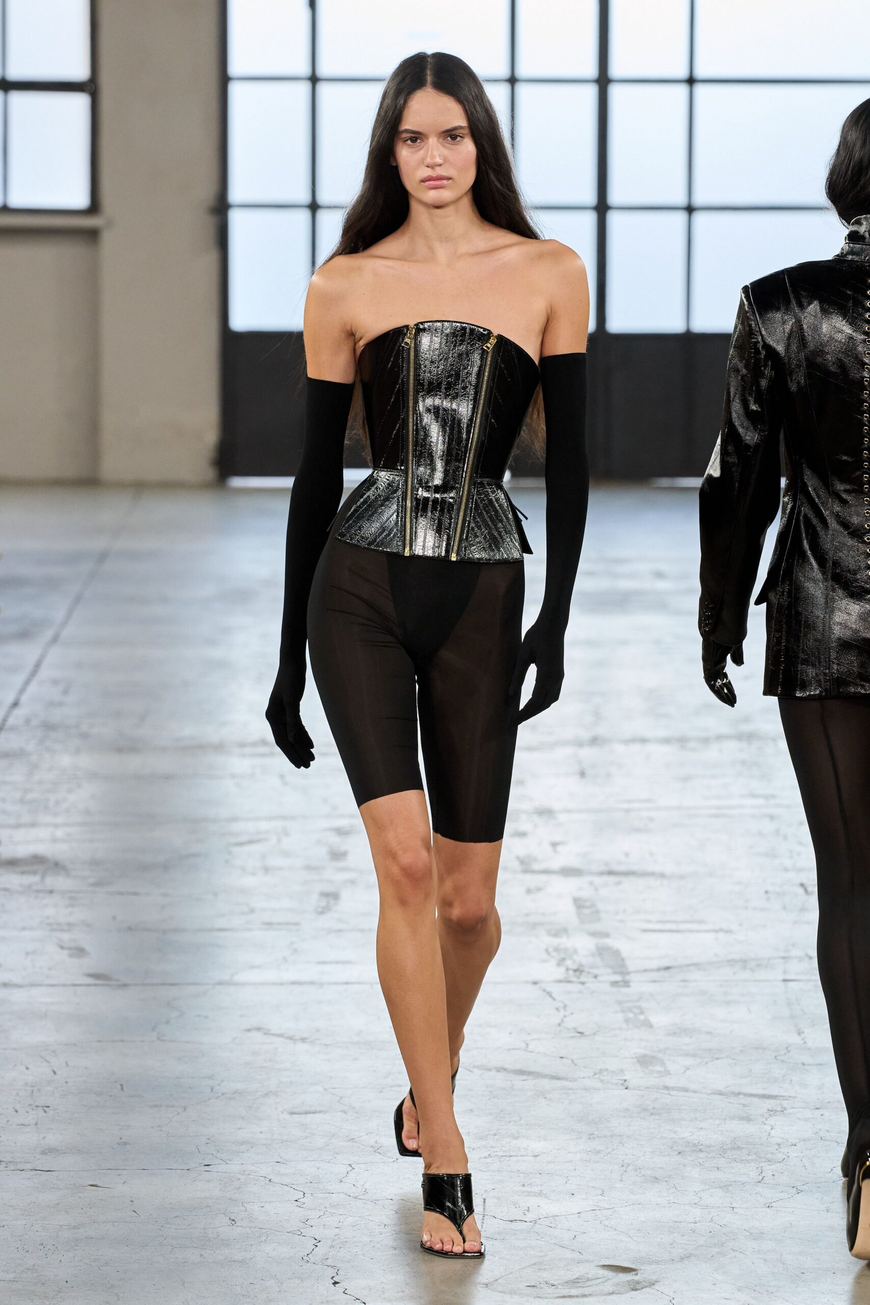 Black patent corset with twin gold zips and a flared peplum. Matte black gloves reaching past the elbow. Sheer black stretch shorts, the high-cut brief visible beneath. Patent thong mules with a narrow kitten heel. Walking across a drafty loft before the afterparty lights come on.