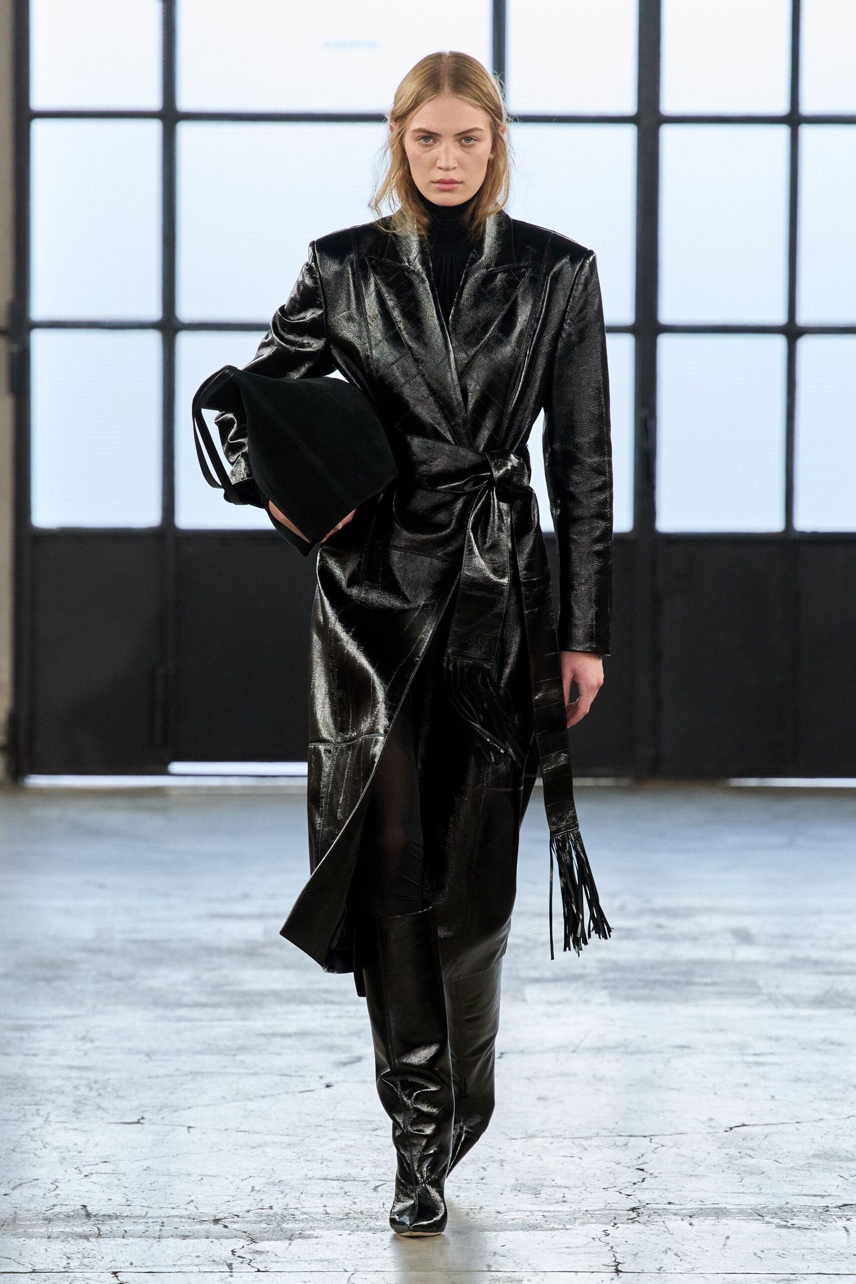 Black glazed leather wrap coat, sharp shoulders and a fringed sash end. Black jersey turtleneck pulled close to the throat. Oversized soft black tote tucked under one arm. Black thigh-high boots in the same glossy leather. Crossing an empty depot office before dawn with the keys.