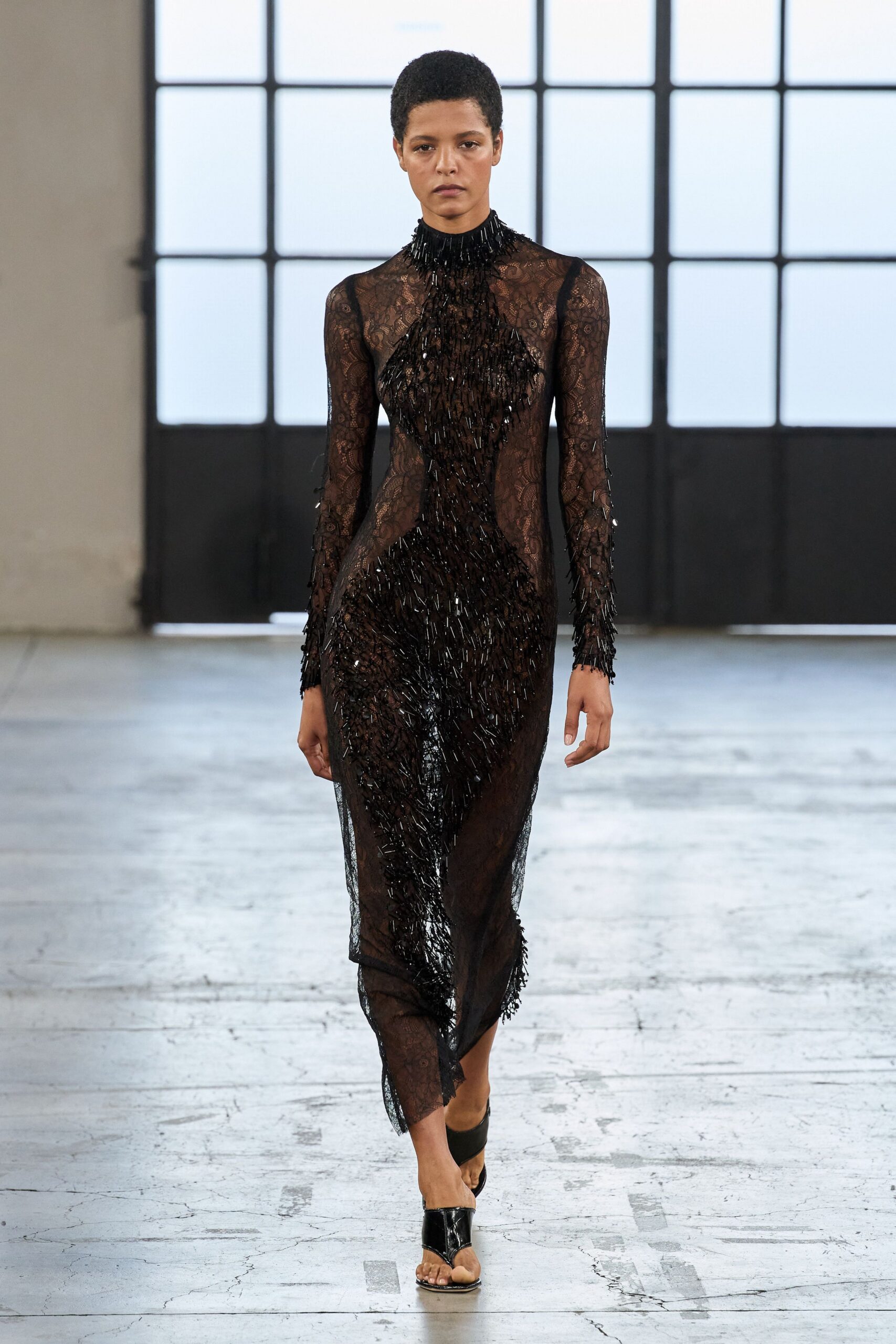 Sheer black lace column gown with bugle-bead fringe thickening into a body line. Black patent thong mules with a sharp squared vamp. After midnight in a drafty ballroom, sequins ground into the floor.