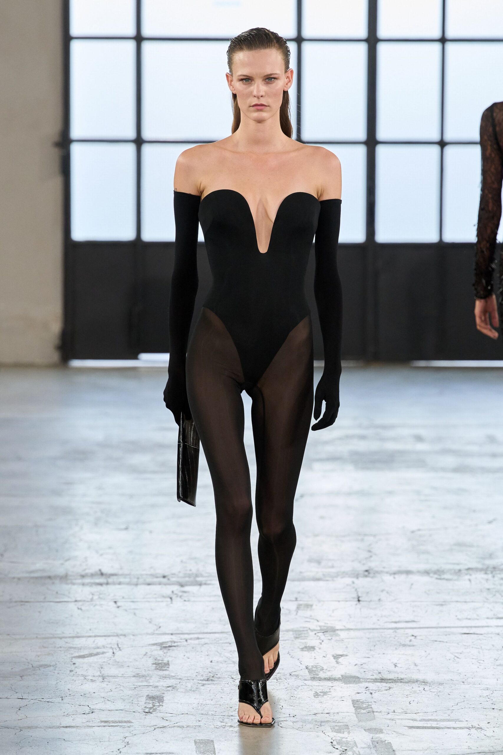 Black stretch bodysuit with a plunging U notch to the sternum. Matching glove sleeves worn off the shoulder. Sheer black tights, denser at the brief line. Croc-embossed black clutch tucked flat at the side. Patent thong sandals with a sharp V over the toes. For crossing a concrete loft when the air-conditioning has given up.