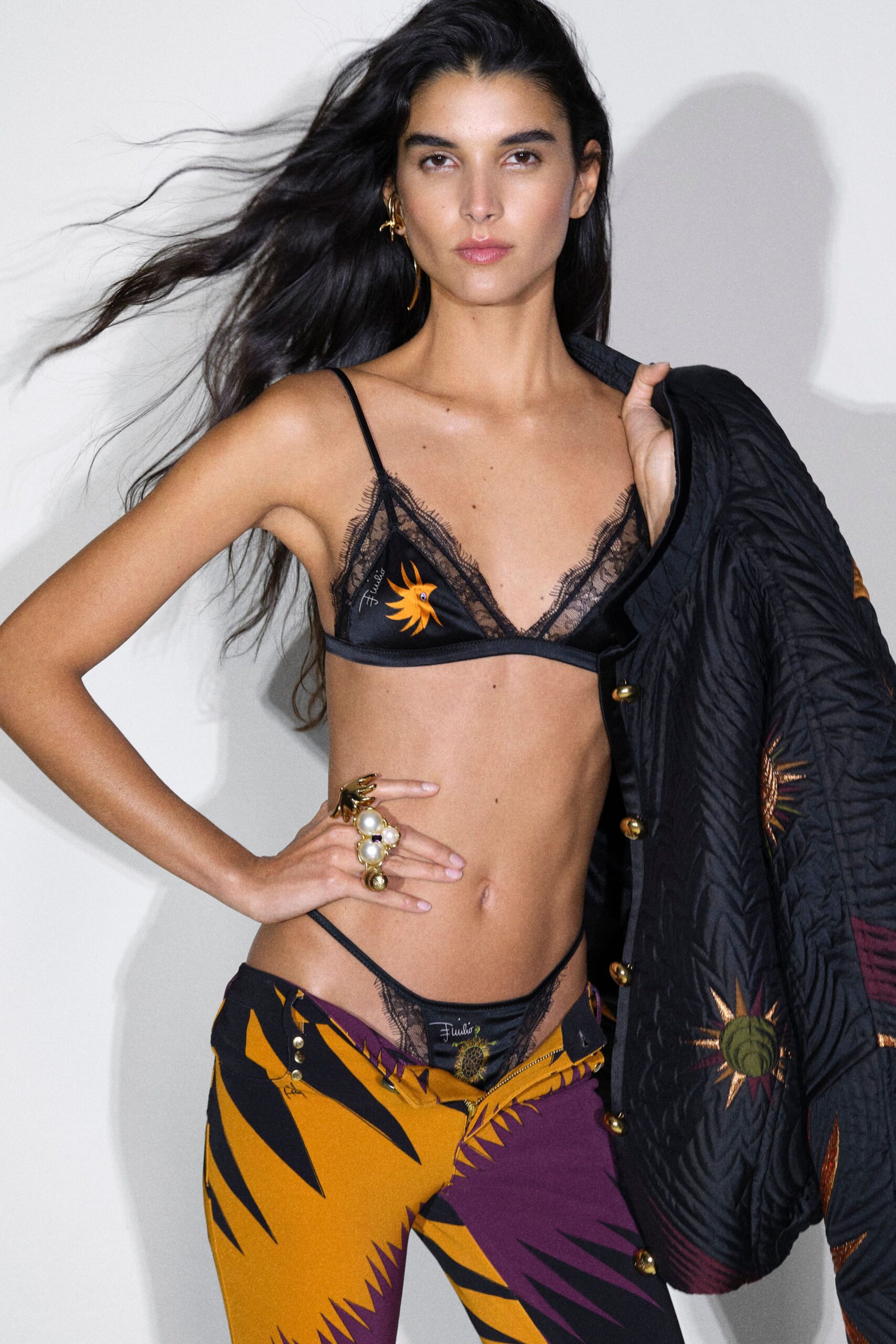 Black satin triangle bra edged in eyelash lace, sunburst motif at one cup. Low-slung trousers in ochre and plum graphic print, zip left half-open. Black quilted jacket with brass snaps over one shoulder. Backstage by the rattling freight lift.