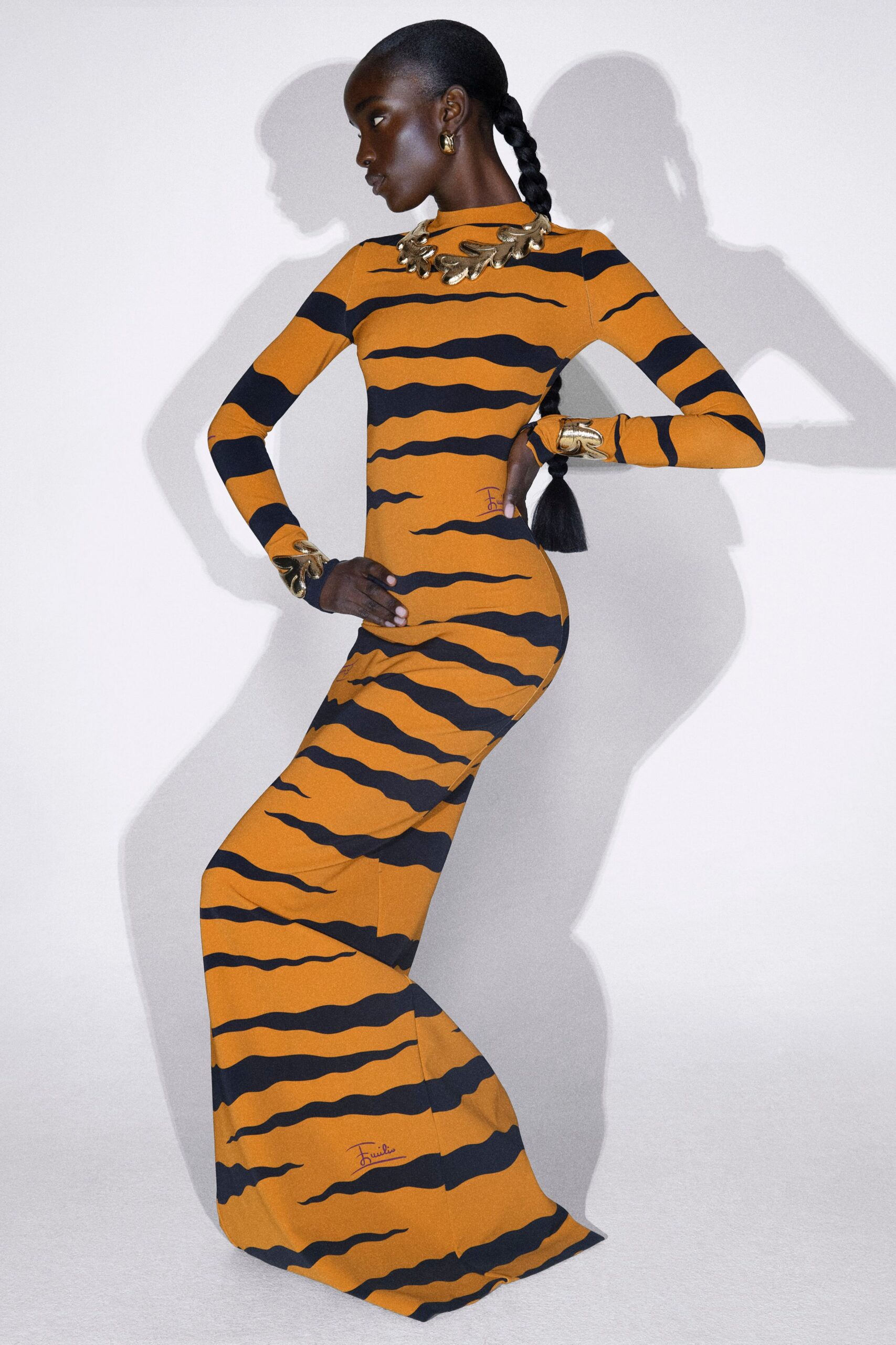 Ochre stretch jersey column dress with a high neck and inky tiger stripes that narrow at the waist, sleeves ending under sculpted gold cuffs. Molten gold collar and hoops. Waiting by the museum wall after everyone else has left.