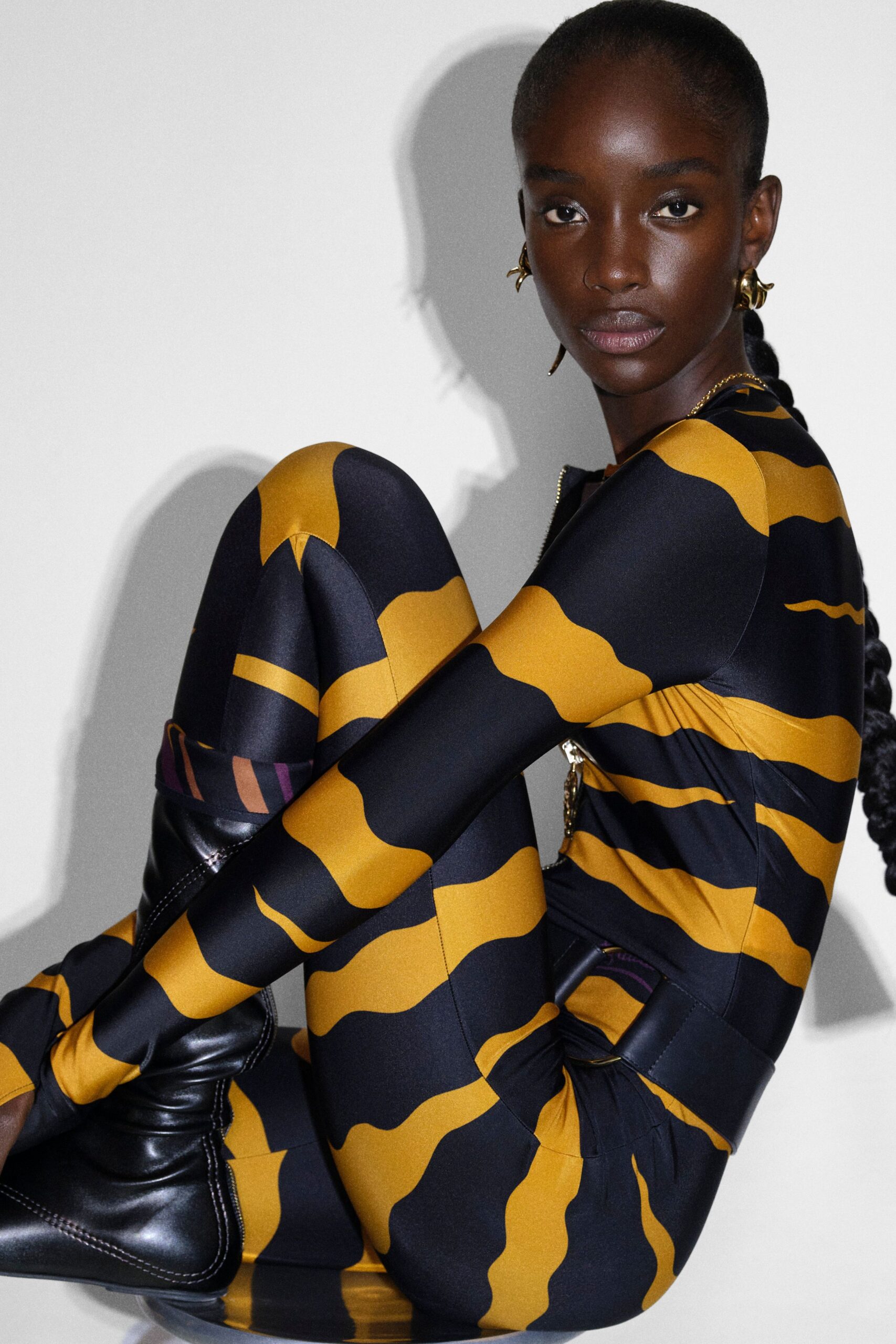 Ochre-and-black stretch catsuit printed in undulating stripes, a tiny front zip glinting at the collar. Black leather knee boots with white topstitching. Waiting in the studio after midnight, shadow doubled on the wall.