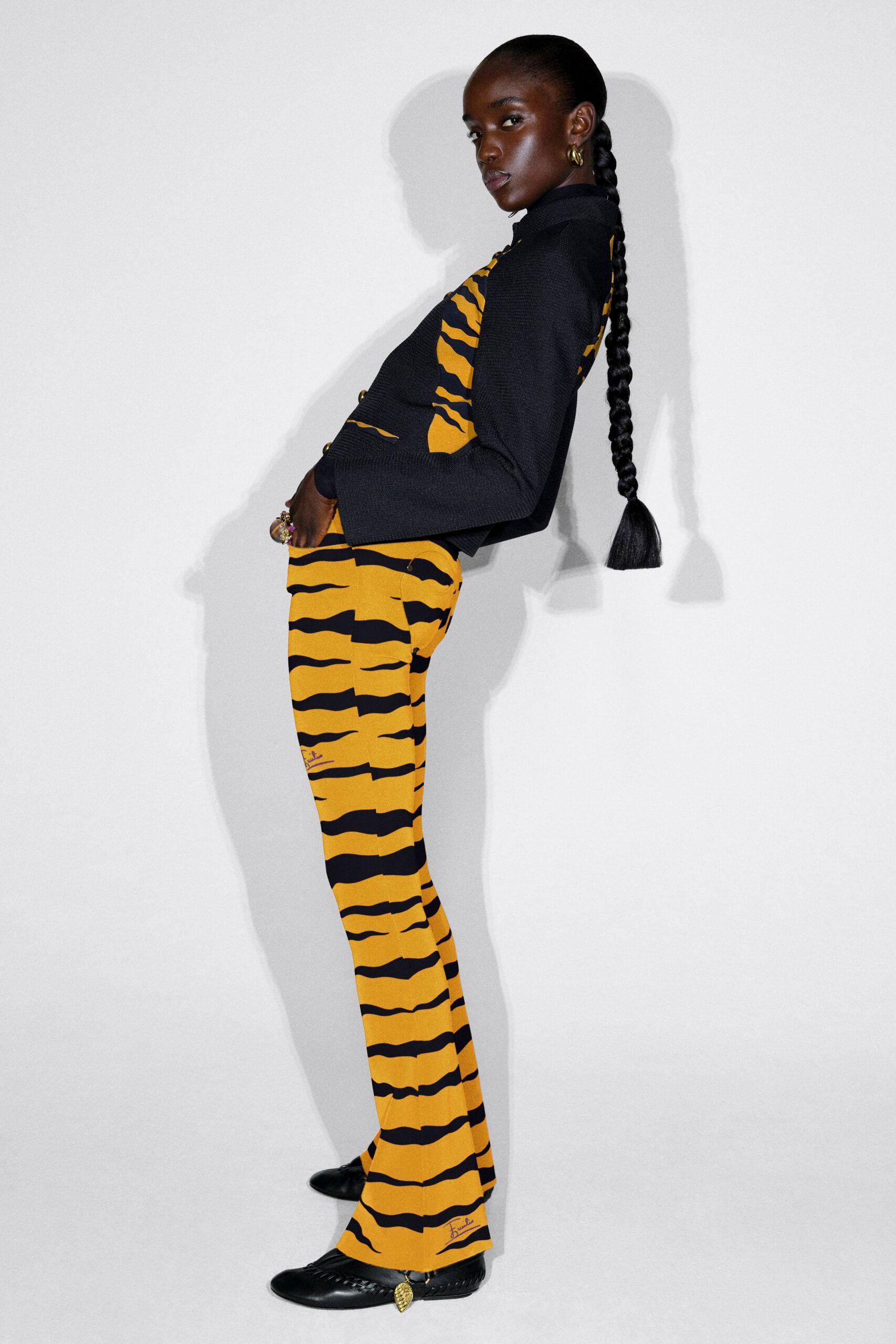 Black waffle-knit jacket with gold buttons over ochre trousers cut in a wavering tiger-stripe print, the hem grazing black woven leather flats with a dangling gold charm. Waiting in the museum corridor after closing.