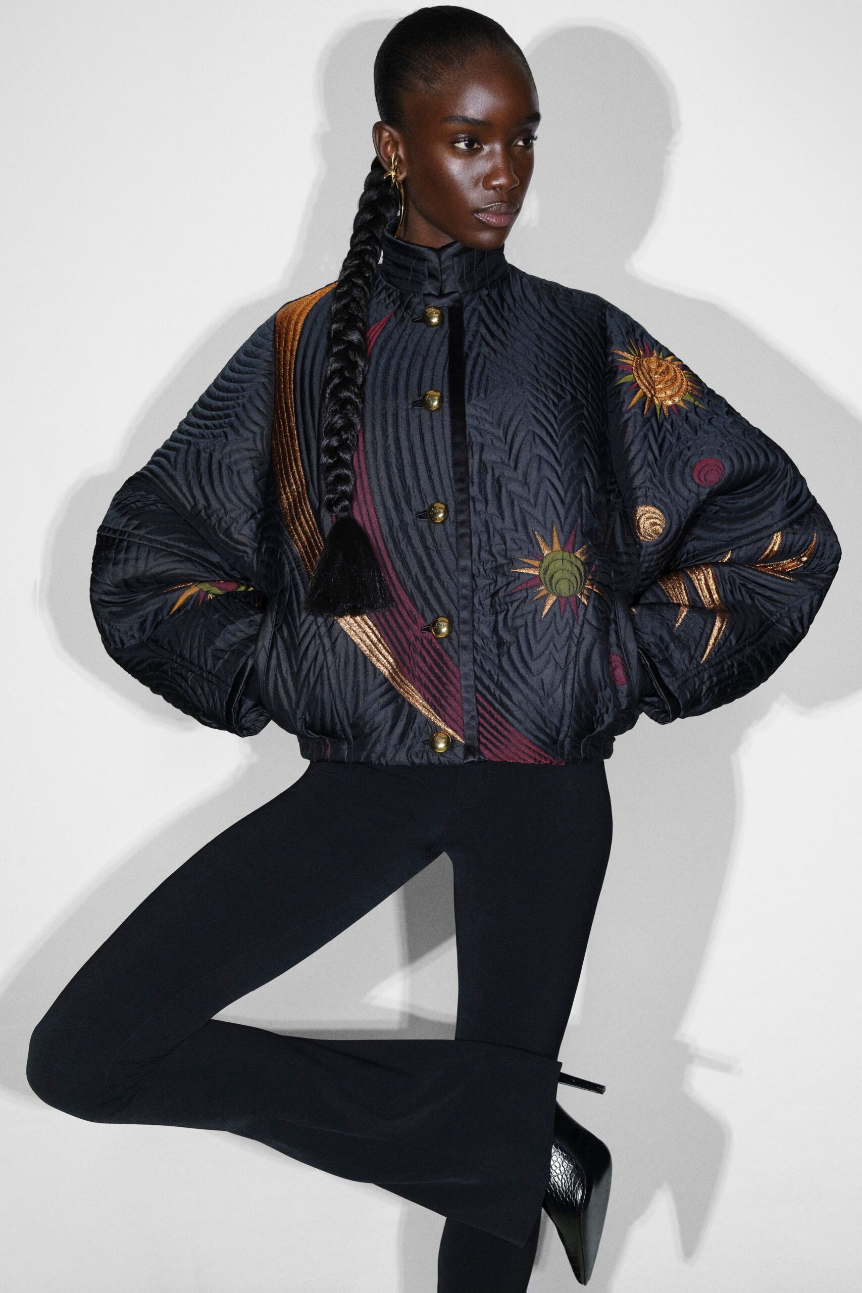 Quilted black jacket with a stand collar, chevron stitching and brass dome buttons, scattered with sunburst embroidery. Black flared trousers, black pointed heels in snakeskin. Waiting in a museum corridor after closing.