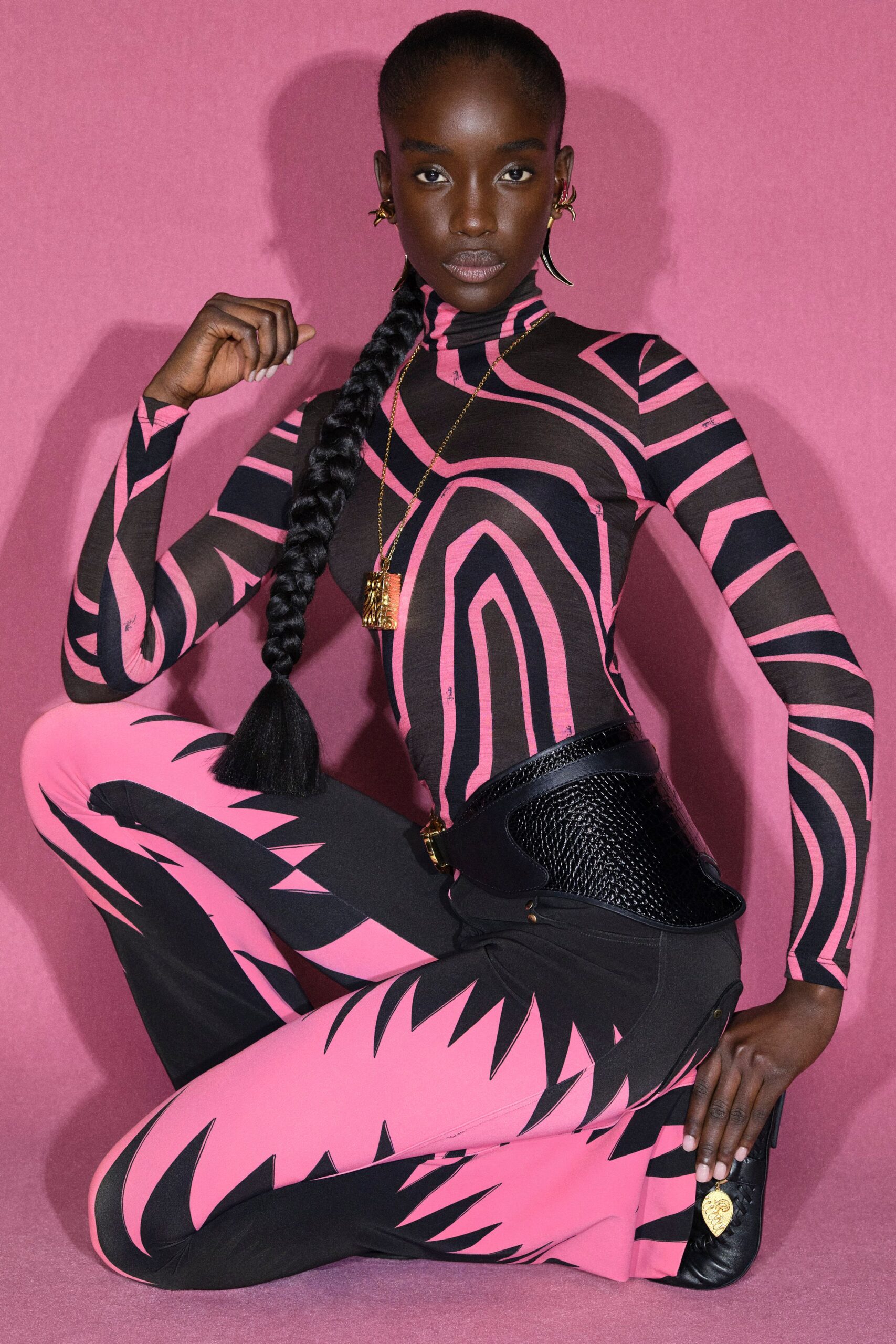 Sheer mesh catsuit in hot pink and black with a close turtleneck and warped stripe print. Black leather corset belt with a basket-woven front panel. Gold pendant necklace. Pink stairwell after midnight, humming fluorescent lights.