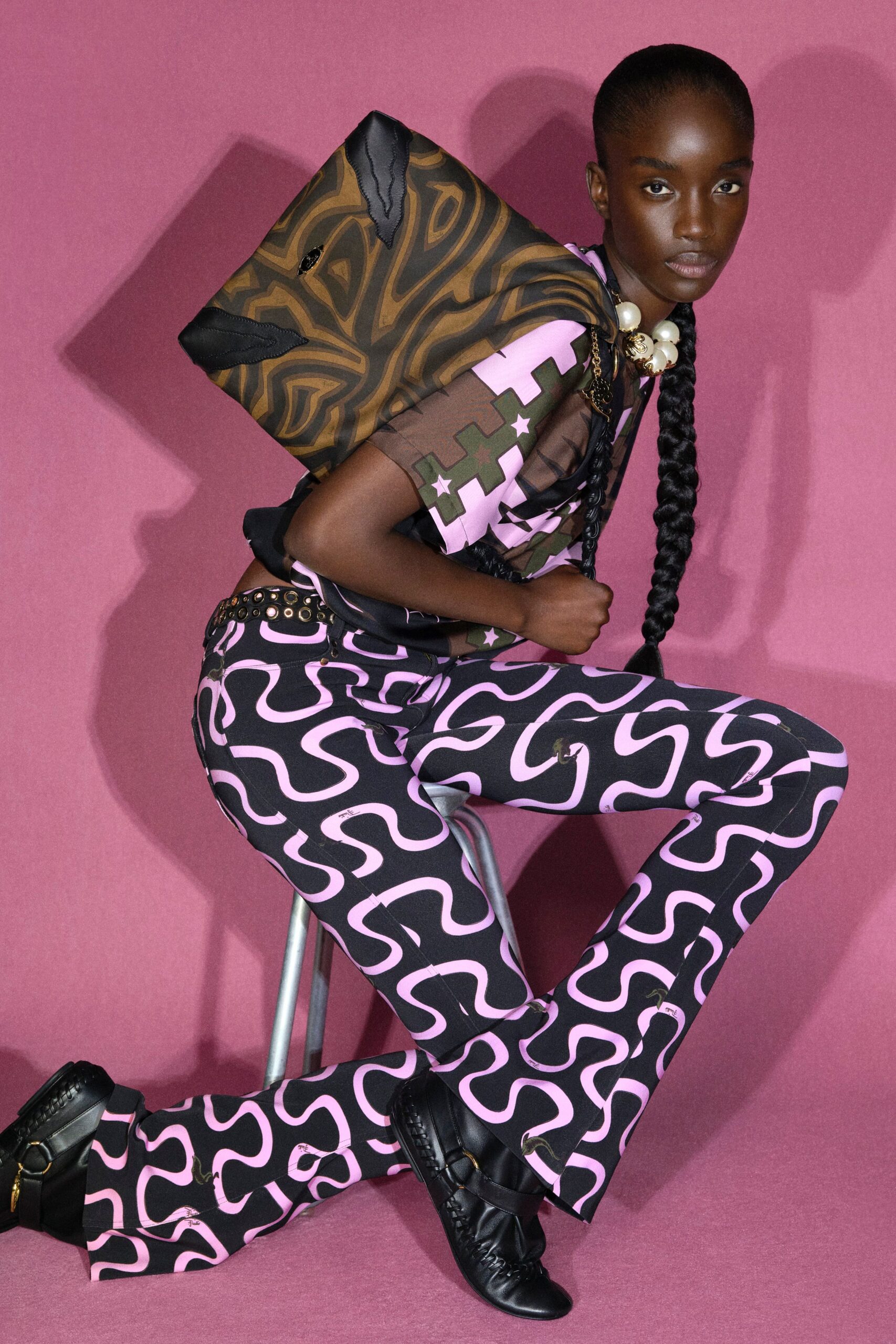 Sheer mesh top spliced with khaki stars and bubblegum puzzle blocks, sleeves ending just below the elbow. Flared trousers in black jersey with pink squiggles, studded belt, black woven leather boots. Perched on a studio stool before the music starts.