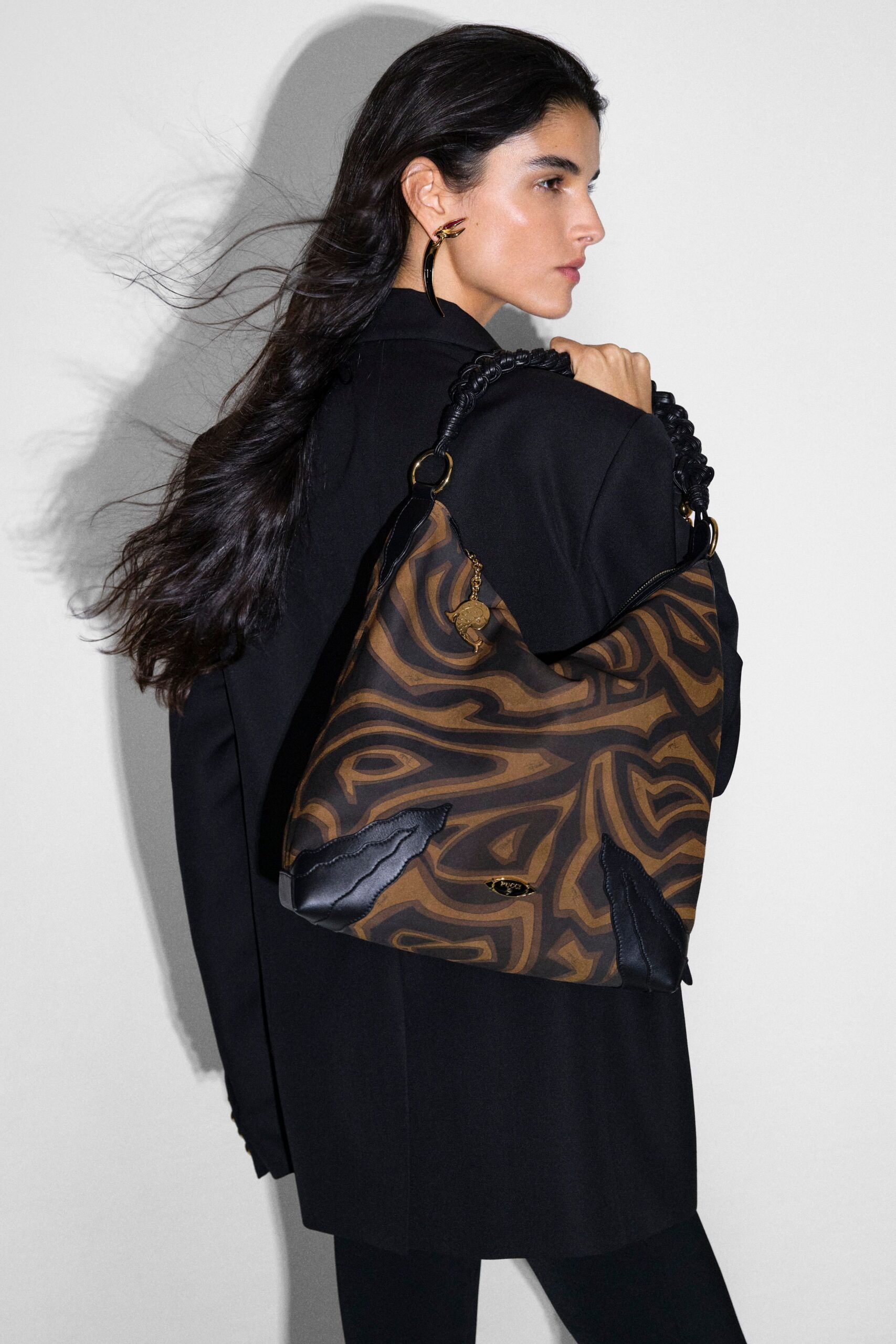 Black tailored coat with a clean vent running down the back, black slim trousers, and a horn-shaped earring in dark enamel. Brown moire-print shoulder bag with braided straps and leather corners. Waiting in a hotel corridor for the lift to arrive.