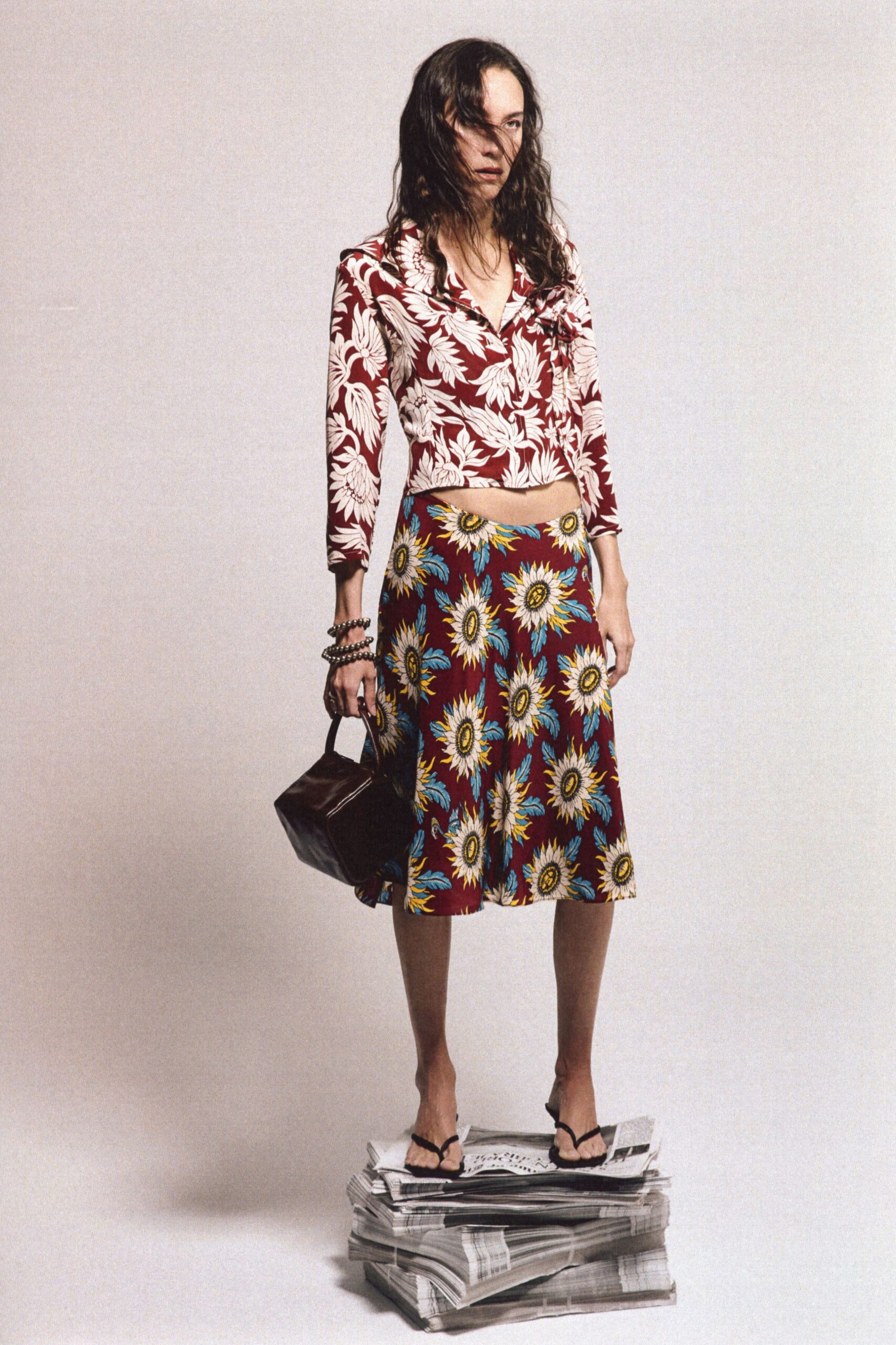 Cropped oxblood jersey top with oversized white hibiscus and a twisted bust knot. A-line midi skirt in the same oxblood, scattered with oversized sunflower heads. Stacked silver bead bracelets. Slouchy black leather box bag. Black thong sandals. Sunday papers underfoot, windows open before the heat starts.