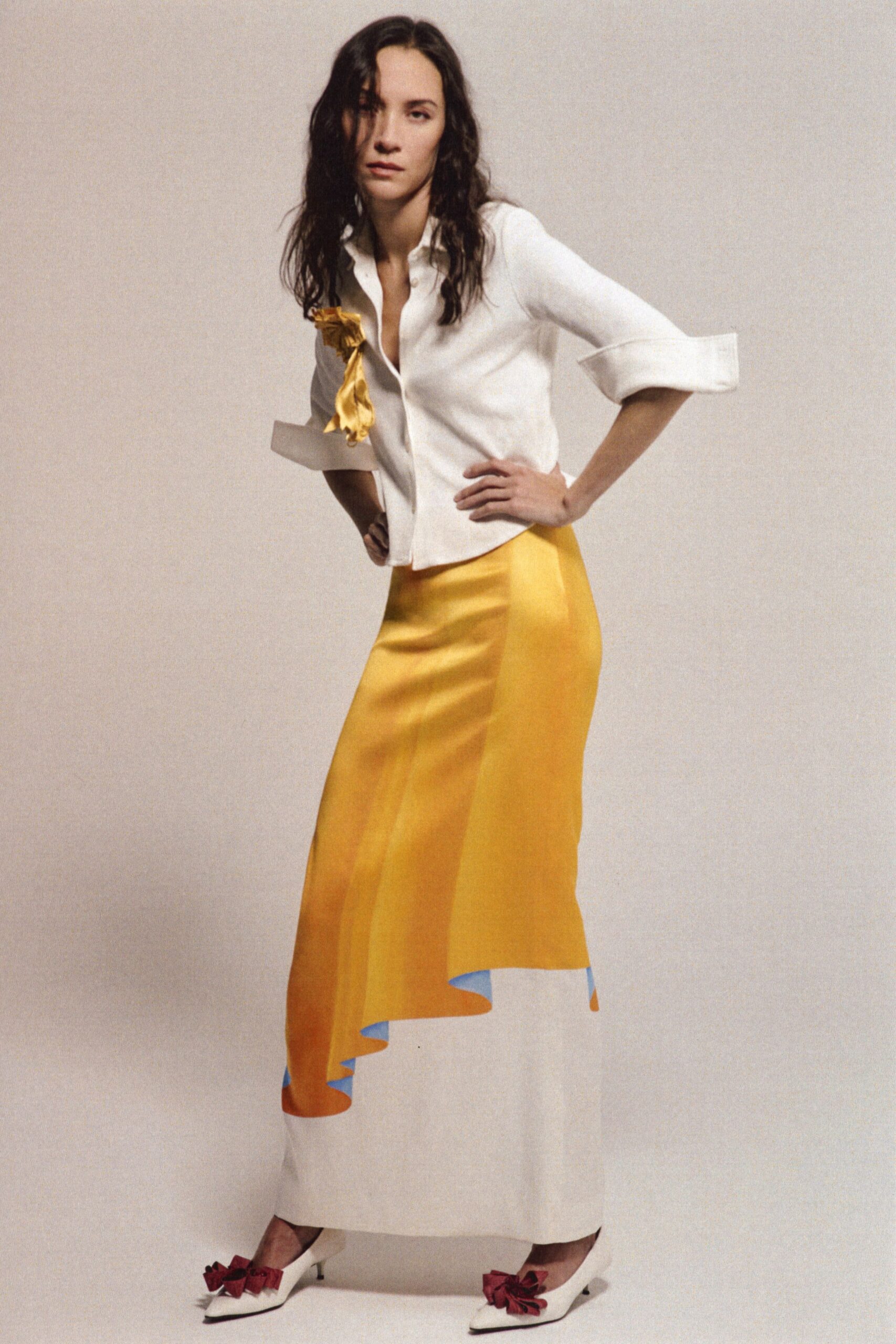 White shirt with deep placket and exaggerated turned-back cuffs. Crushed gold satin bow pinned high on the chest. Bias-cut satin skirt in marigold fading to white, hem undulating blue underneath. White pointed pumps topped with dark red rosettes. Hotel ballroom at noon, flowers already dropping from the centerpieces.