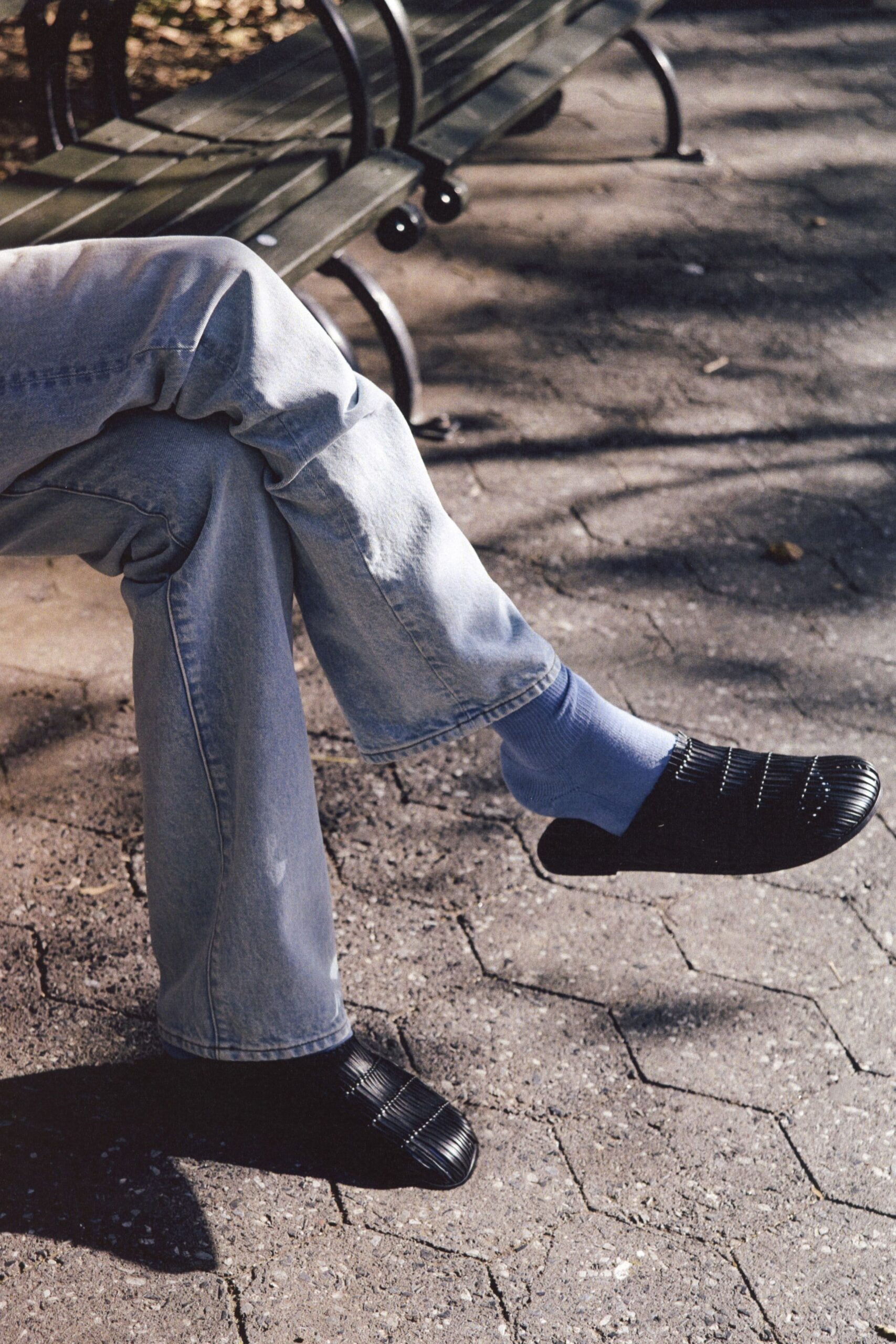 Pale stonewashed denim jeans, cut wide with a soft break at the hem. Powder-blue ribbed socks pulled high above the shoe line. Black flats with a glossy ridged cap and squared-off toe. Sitting on a park bench after lunch, sun shifting through the trees.