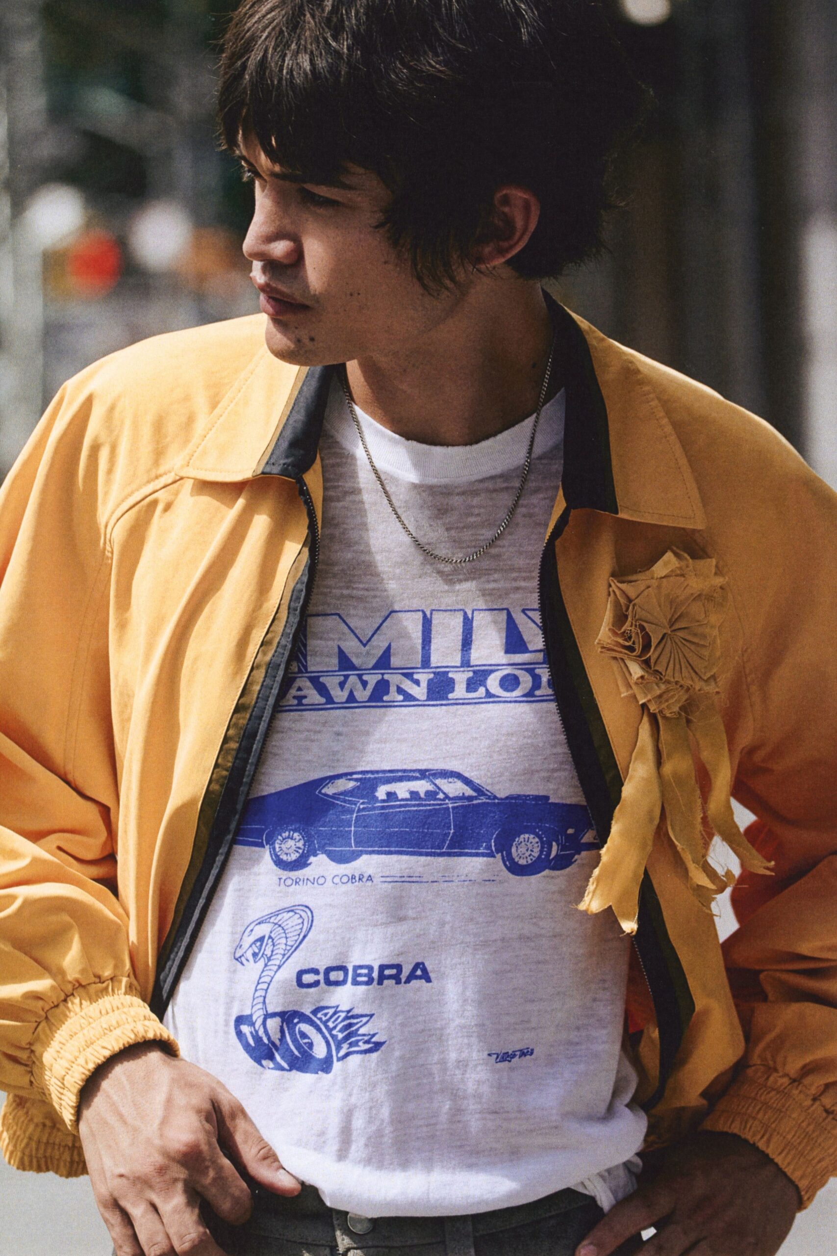 Buttermilk zip jacket with black facings and a crumpled rosette pinned askew. White marl T-shirt printed in cobalt with muscle car and cobra graphics. Fine silver curb chain at the collarbone. Leaning on a side street railing after buying a coffee and cigarettes.