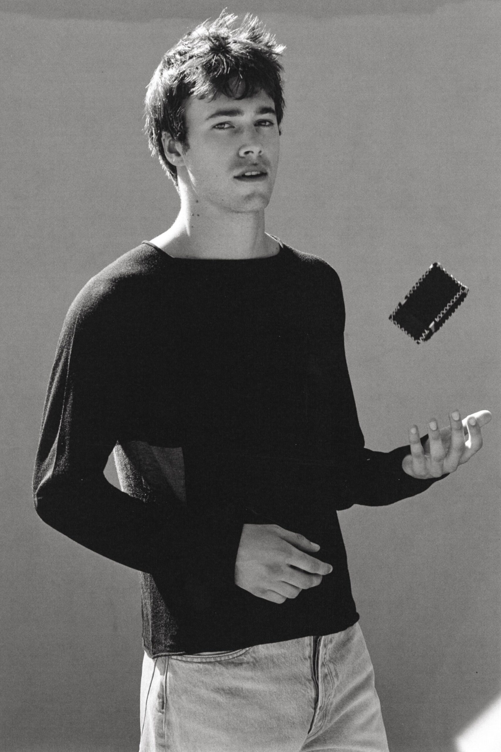 Fine dark knit sweater with a wide boatneck and pale side inset. Light-wash jeans, zip fly centered stark against the washed denim. Small rectangular wallet with pinked edges tossed above the hand. Waiting outside the corner shop, killing time before sunset.