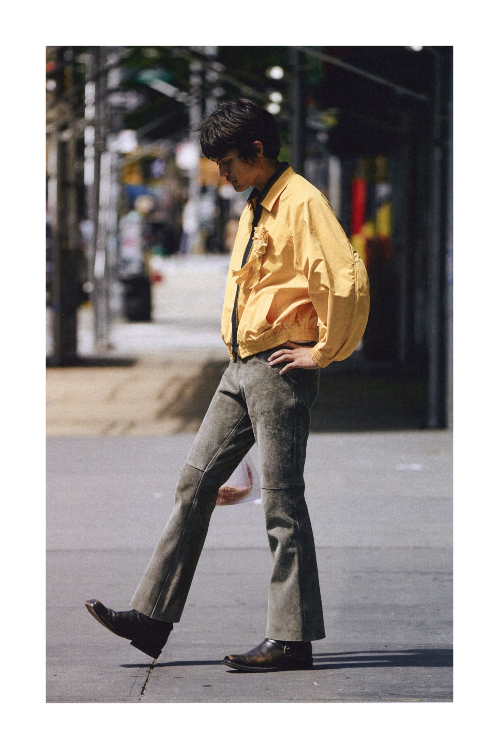Butter-yellow cotton blouson, elastic hem ballooning under a black zip. Dark underlayer peeking at the collar and placket. Moss-grey suede flares with knee seams and a dusty nap. Dark leather ankle boots with a low stacked heel. Crossing the street after buying fruit, before the heat leaves the pavement.