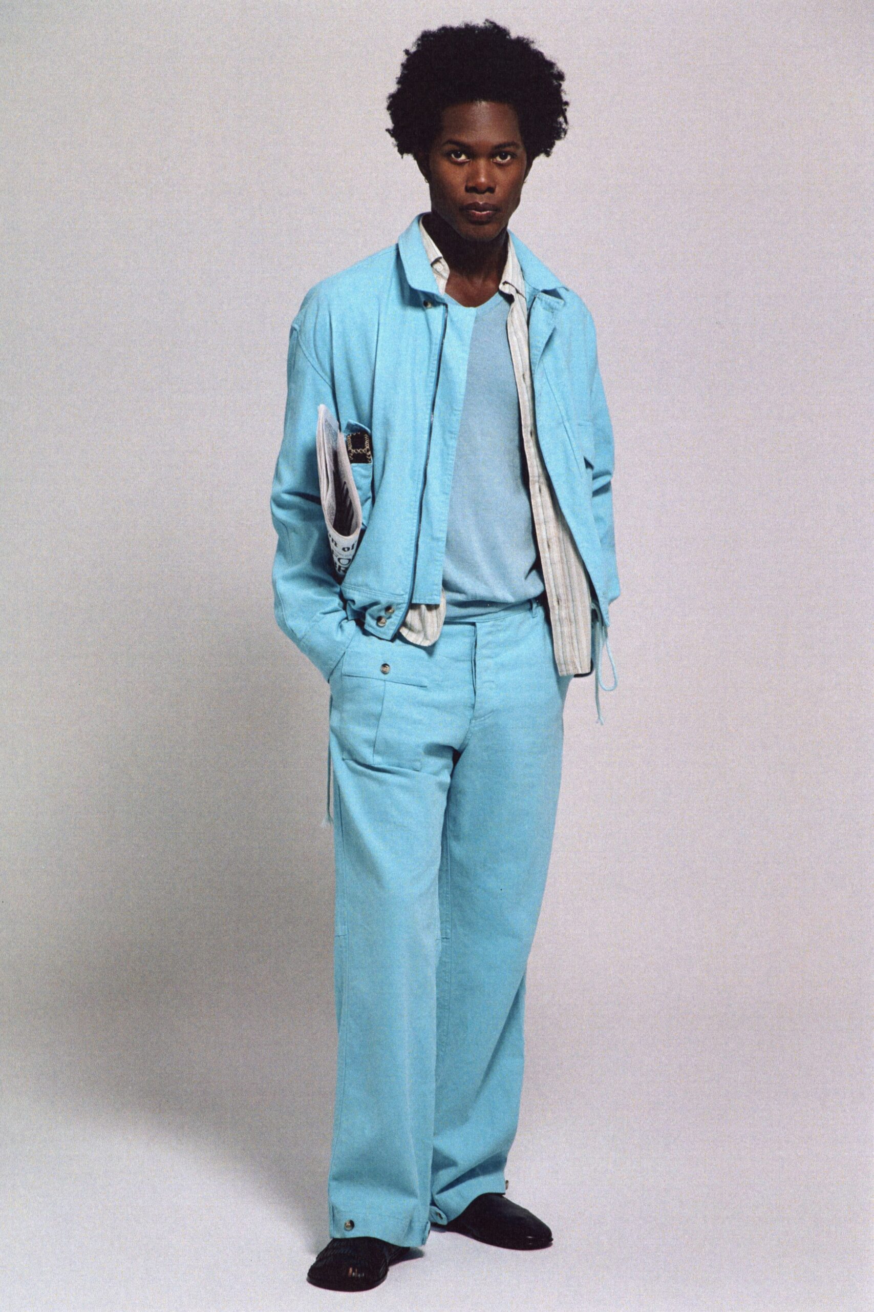 Powder-blue jacket with a short hem and dangling side drawcord. White striped shirt hanging open under the collar. Fine-gauge sky knit, cut plain and close. Matching wide trousers with flap pockets and snapped cuffs. Black square-toe slip-ons, lightly ribbed. Walking into a seaside hotel lobby before anyone has checked in.