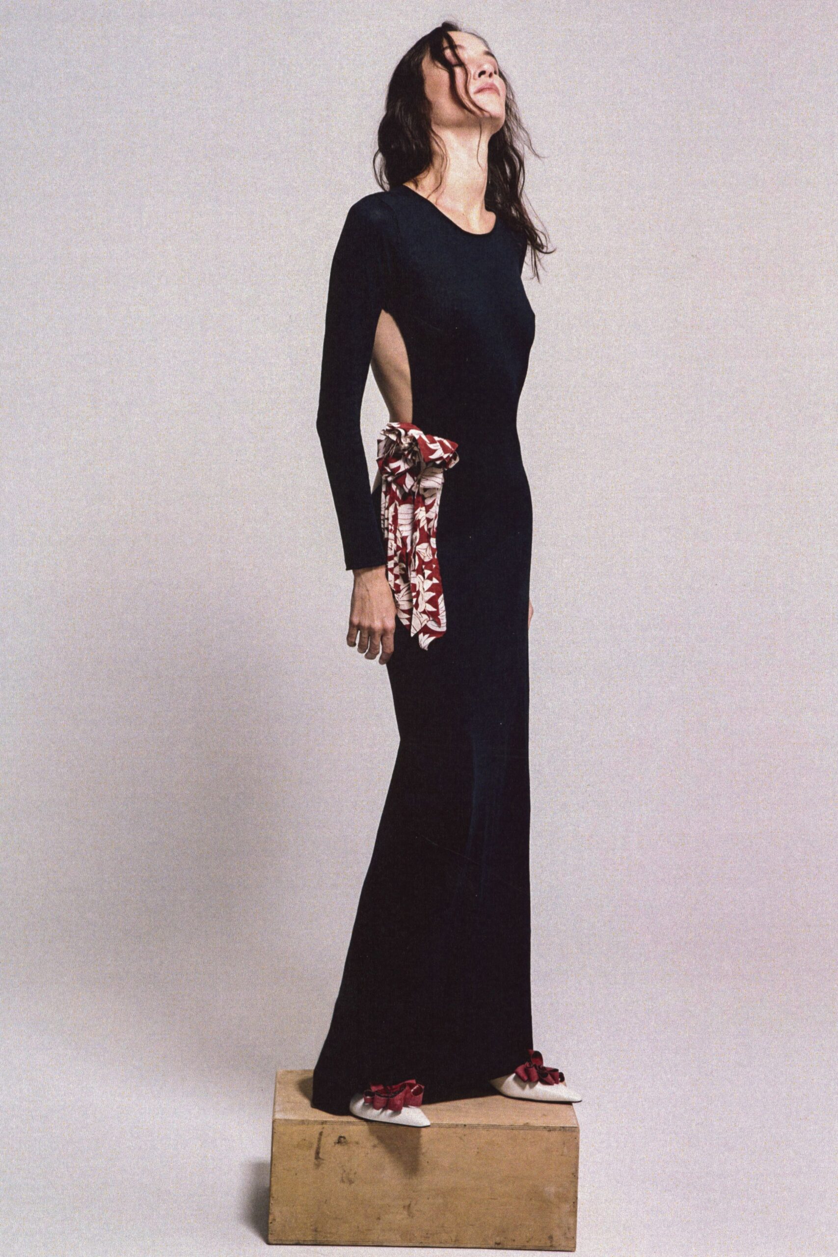 Black jersey column gown with a deep side cutout and long sleeves. Rust-and-cream floral sash knotted high at the waist, ends hanging loose. Pointed ivory flats topped with crushed oxblood bows. For crossing a tiled courtyard after the wedding dinner has thinned out.