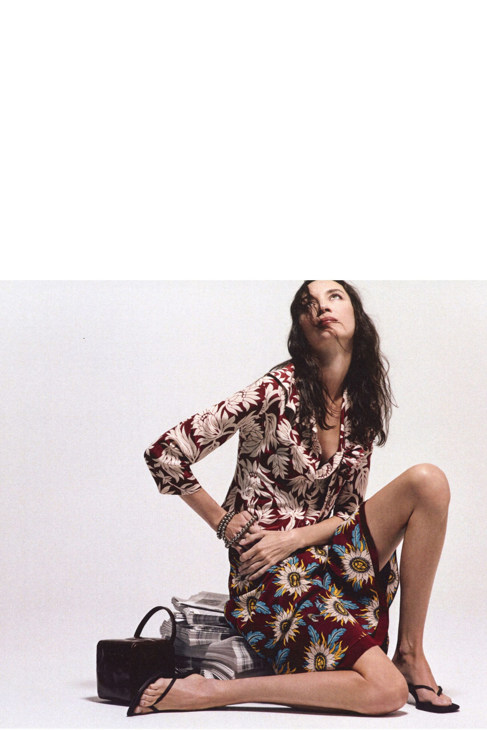 Burgundy blouse in cream tropical florals, plunging neckline softly twisted at the chest. Matching wrap skirt printed with oversized daisies, slit opening high on the thigh. Stacked gunmetal bead strands at one wrist. Black thong sandals with flat soles. Sunday papers on the floor, windows open before the heat turns serious.