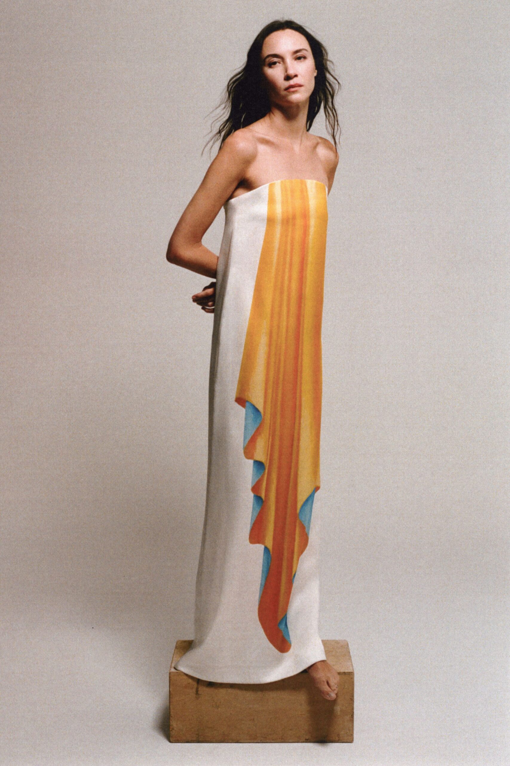 Strapless white column gown with a painted saffron panel falling in scalloped tiers, sky-blue underside flashing at each curve. Standing barefoot on a packing crate while the wall paint is still drying.