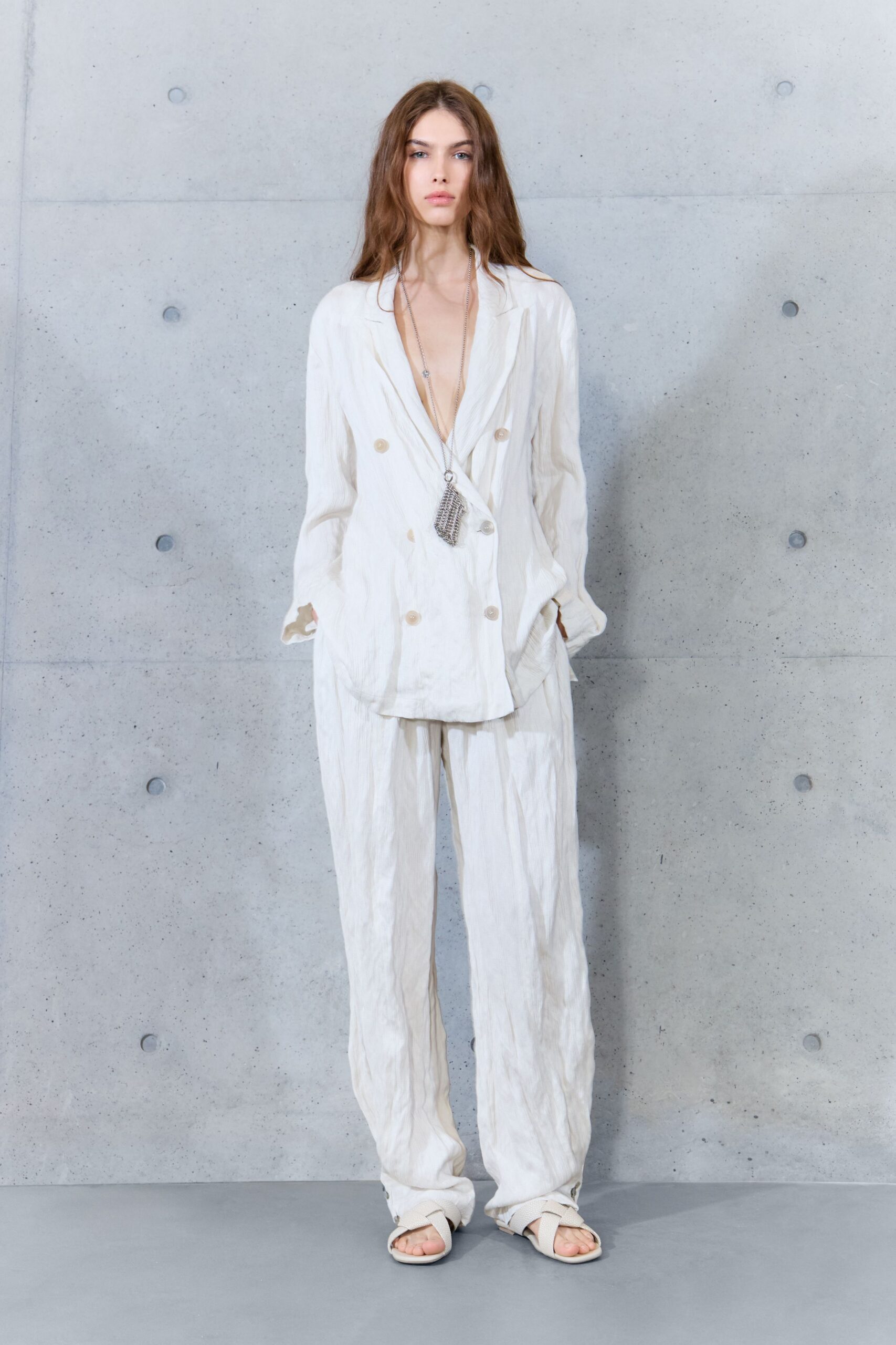 Ivory crinkled blazer, double-breasted but worn plunging almost to the waist. Long silver chain ending in a tiny mesh pouch. Loose ivory trousers in the same crushed cloth, pooling over the feet. Pale crossover sandals with broad matte straps. Hotel terrace at breakfast, sea air already curling the table linen.