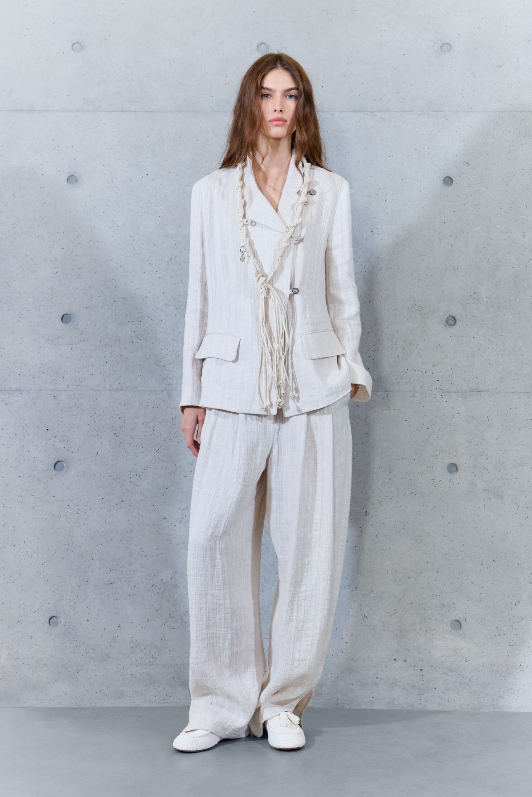 Chalk crinkled jacket with offset buttons and a looped fringe tie. Loose matching trousers in airy gauze, pooling over the instep. White woven slip-ons, one lace end dragging forward. For the ferry crossing when the island is still half asleep.