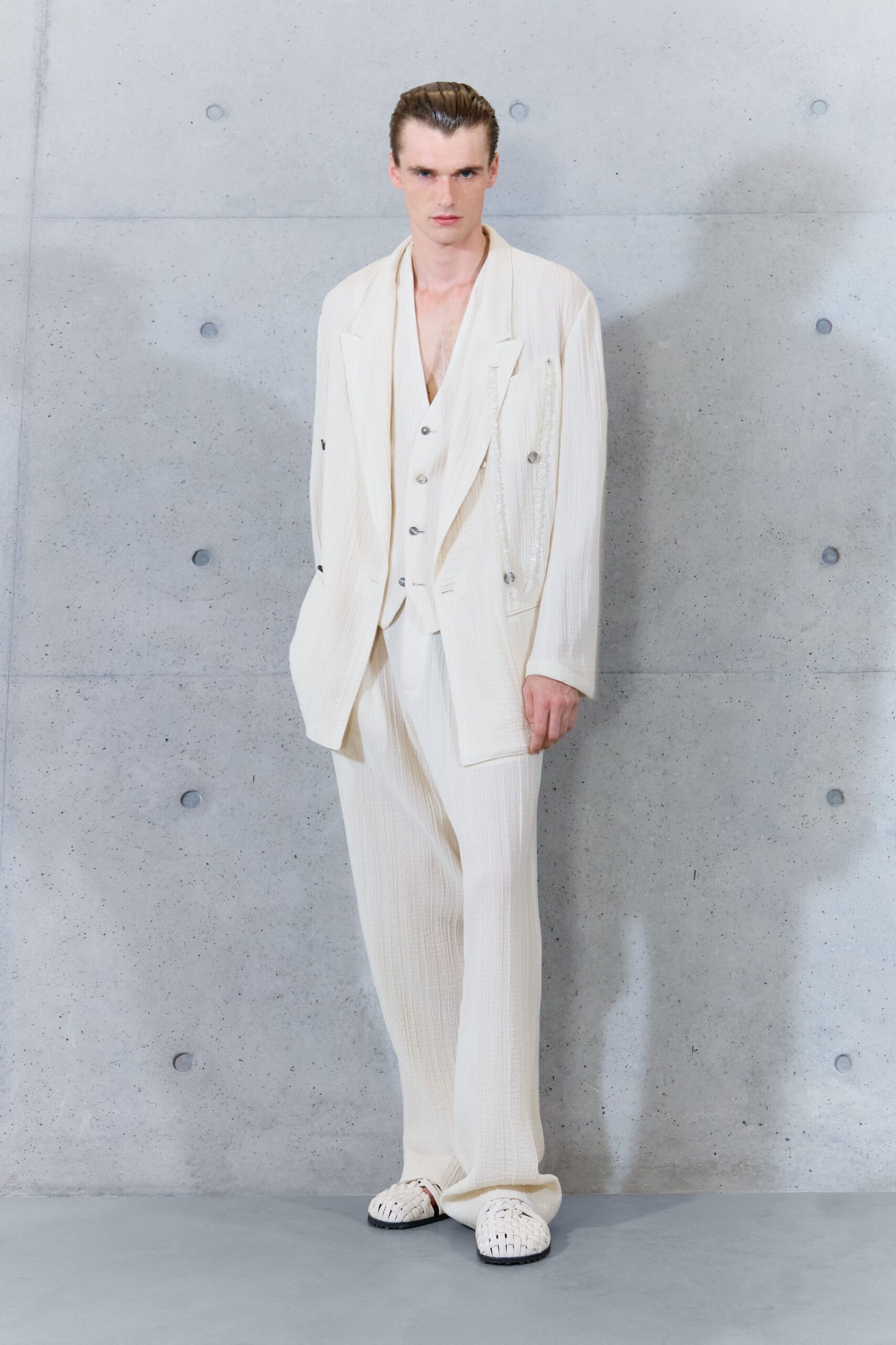 Ivory crinkled blazer with slouched lapels and a frayed vertical inset. Deep-V matching vest fastened with small pewter buttons. Loose ivory trousers in the same puckered weave, pooling at the hem. White woven leather fisherman sandals with black tread soles. Sunday civil ceremony in a concrete chapel, flowers still in buckets.