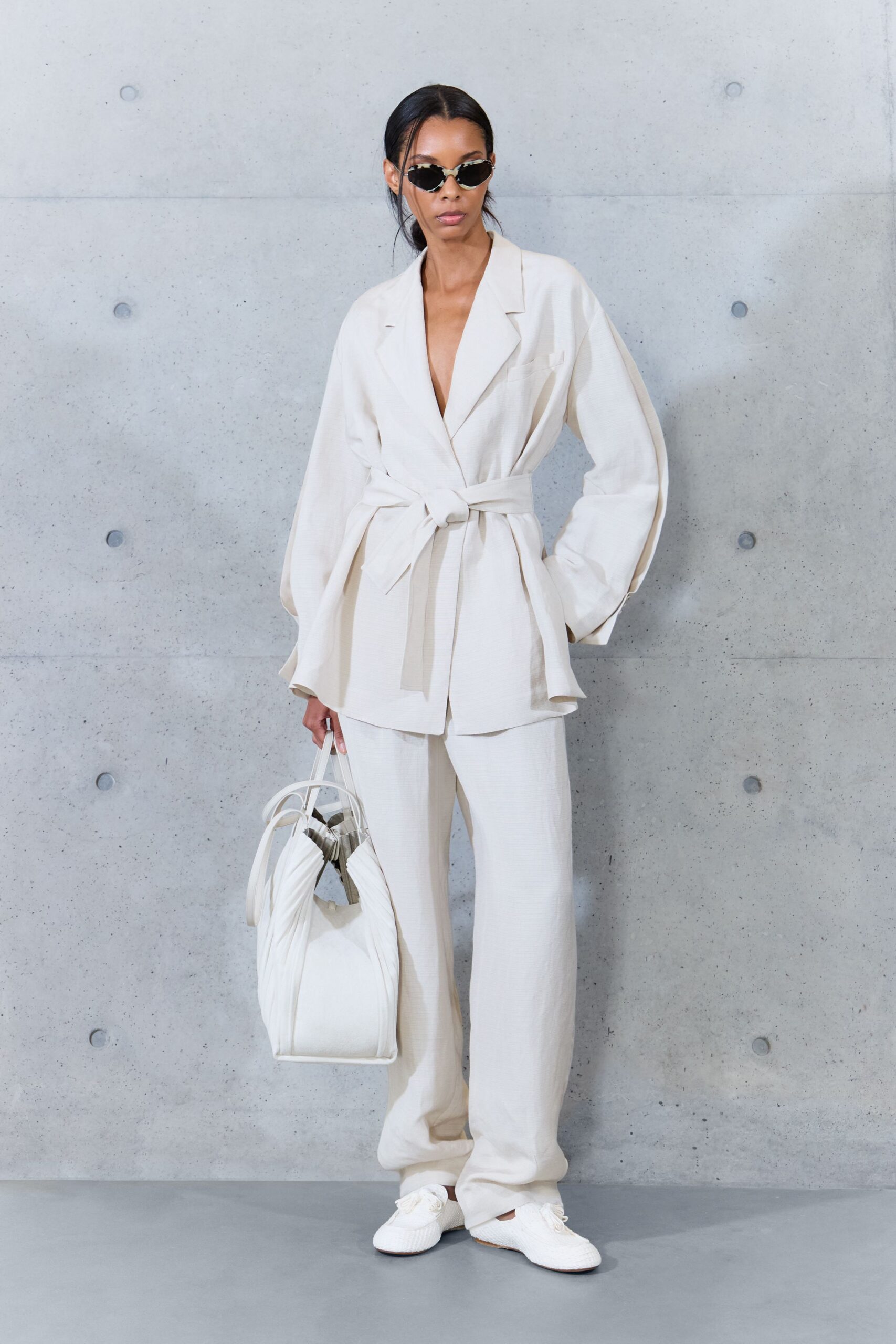 Tiny black cat-eye sunglasses with silvered rims. Ivory wrap jacket in dry crepe, tied low with a thick sash. Matching wide trousers, pooling softly over the instep. White gathered bucket bag with looped handles and mirrored lining. White woven lace-up flats. Early check-in at a concrete hotel, luggage already sent upstairs.