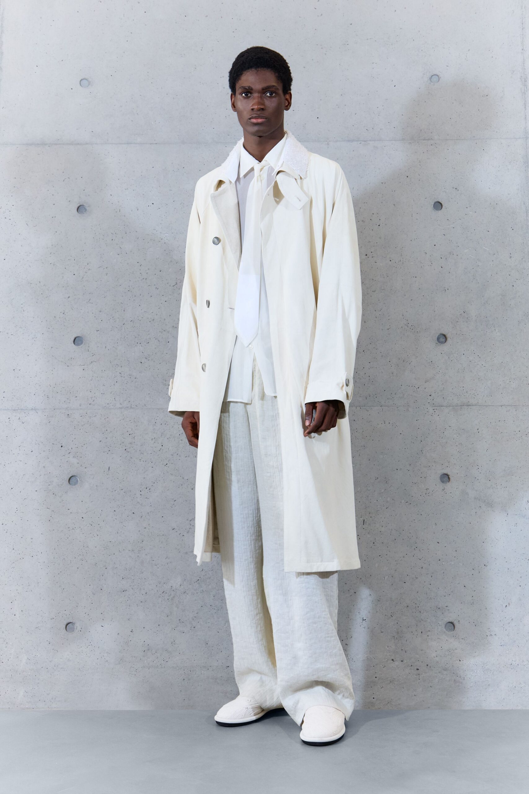 Chalk trench in crisp cotton, silver buttons and loose throat latch. White shirt with a long narrow tie panel falling straight down. Wide ivory trousers in slubby linen, breaking softly over the shoes. Quilted white slip-ons with rounded toes. Early ferry departure, sea fog still sitting on the railings.