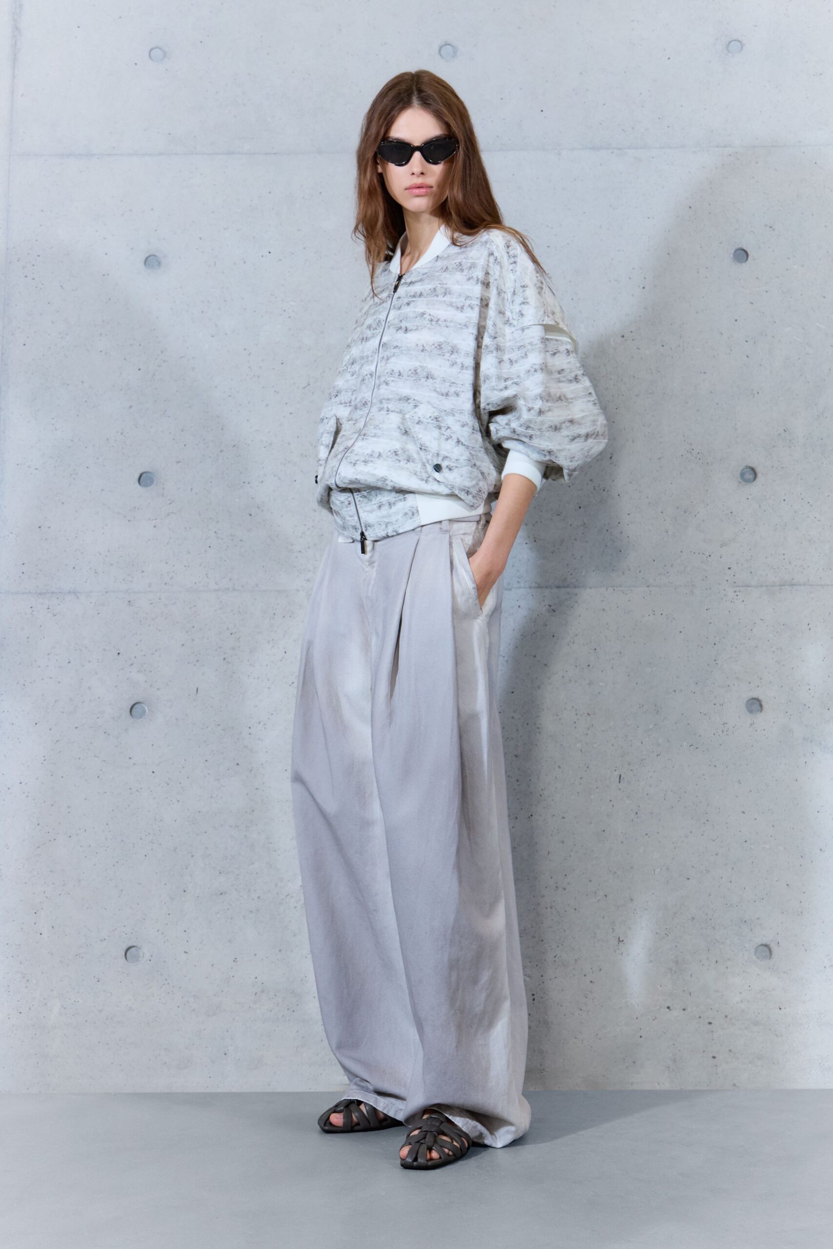 Black cat-eye sunglasses with narrow glossy frames. Clouded grey-and-white bomber, zip ending at a ribbed white hem. Pearl-grey satin trousers cut extra wide with deep front pleats. Black woven leather sandals. Early ferry crossing, concrete still cold underfoot.