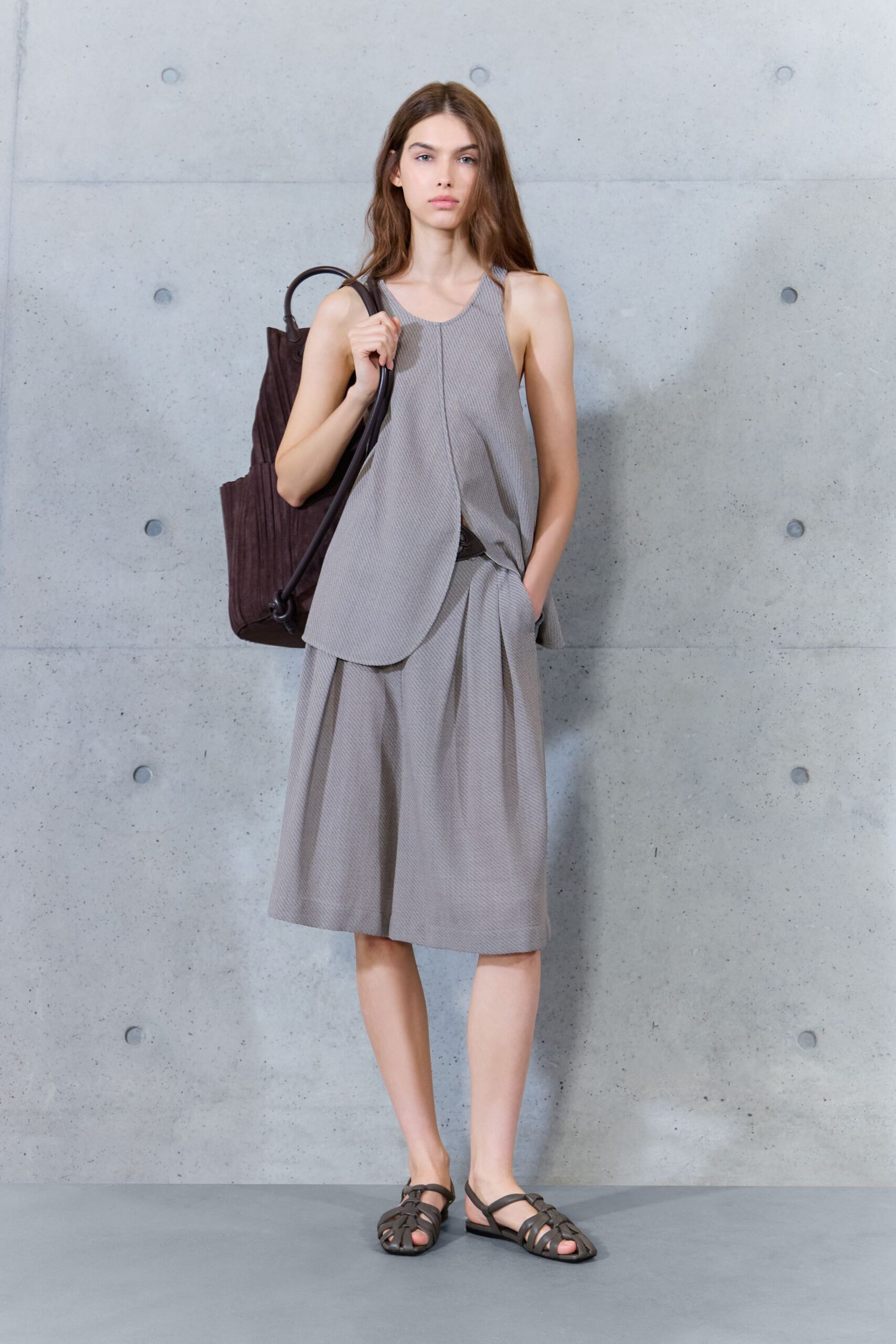 Greige micro-textured tank, front panel curving into a long overlap. Matching calf-length culottes with deep inverted pleats and side pockets. Dark chocolate suede tote with a looped leather shoulder strap. Brown fisherman sandals, flat-soled. Walking across a quiet museum plaza before the heat lifts off the concrete.