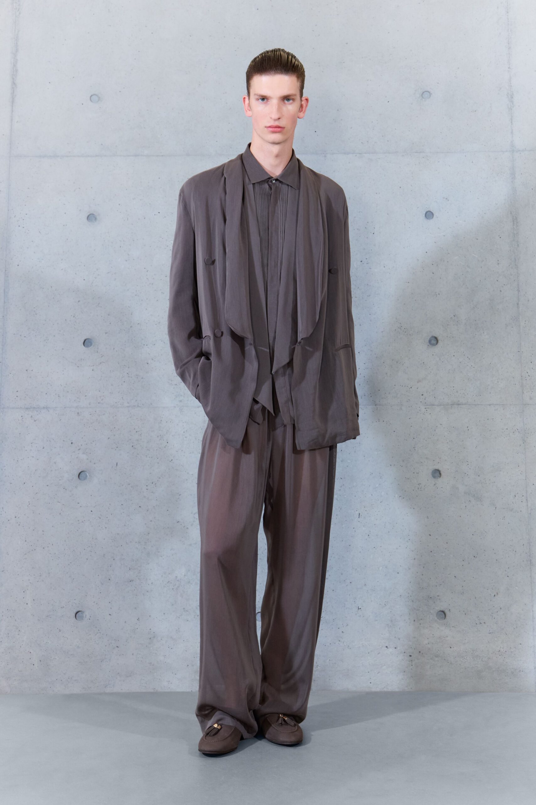 Taupe fluid jacket with hanging scarf panels and covered side buttons. Matching shirt with narrow pleats running from collar to placket. Loose straight trousers in the same drapey cloth, breaking over the shoe. Soft taupe loafers with a small gold horsebit. Waiting in a concrete lobby before the architect comes down.