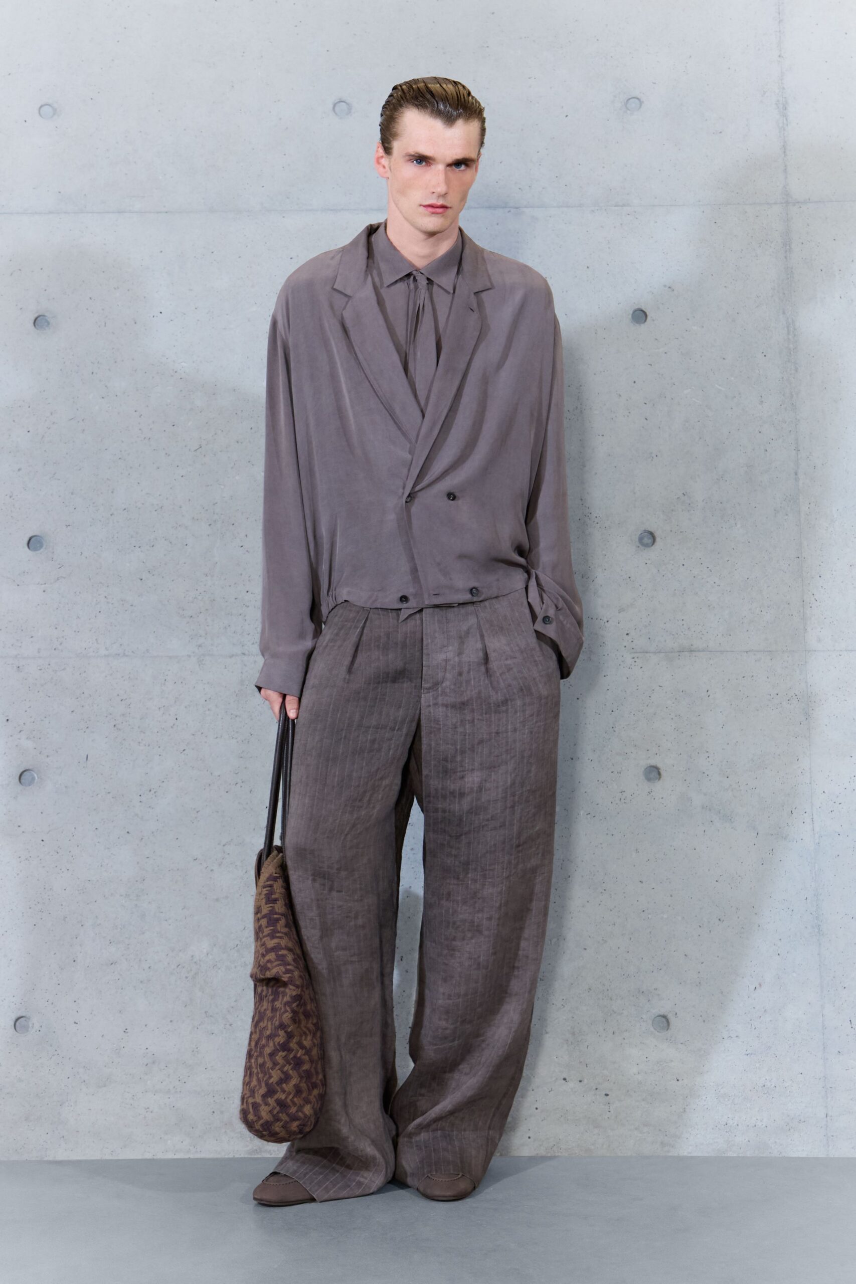Taupe shirt with a narrow self-tie dropping from the collar. Fluid taupe blazer, cut low with one skewed button left visible. Loose brown pinstripe trousers, double pleats breaking over the shoe. Long hobo bag in brown chevron weave. Square-toe taupe slip-ons. For the quiet hour in a hotel lobby before anyone else comes down.