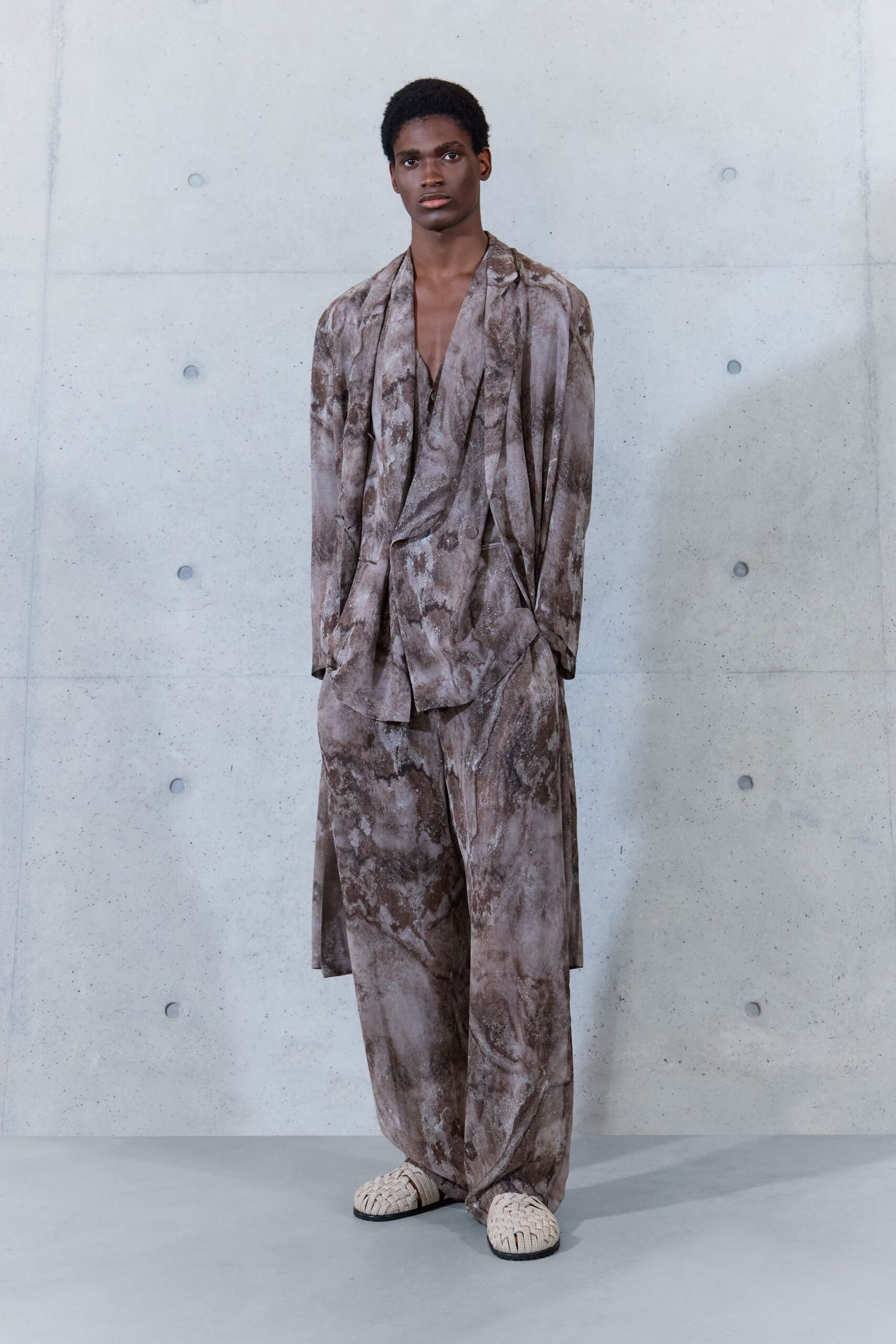 Dusty taupe washed jacket, lapels falling open in soft uneven folds. Matching draped shirt, deep V opening and a low third button. Wide trousers in the same clouded print, pooling over the instep. Cream woven leather slippers with black soles. Concrete atrium at noon, carrying yesterday's heat in the walls.