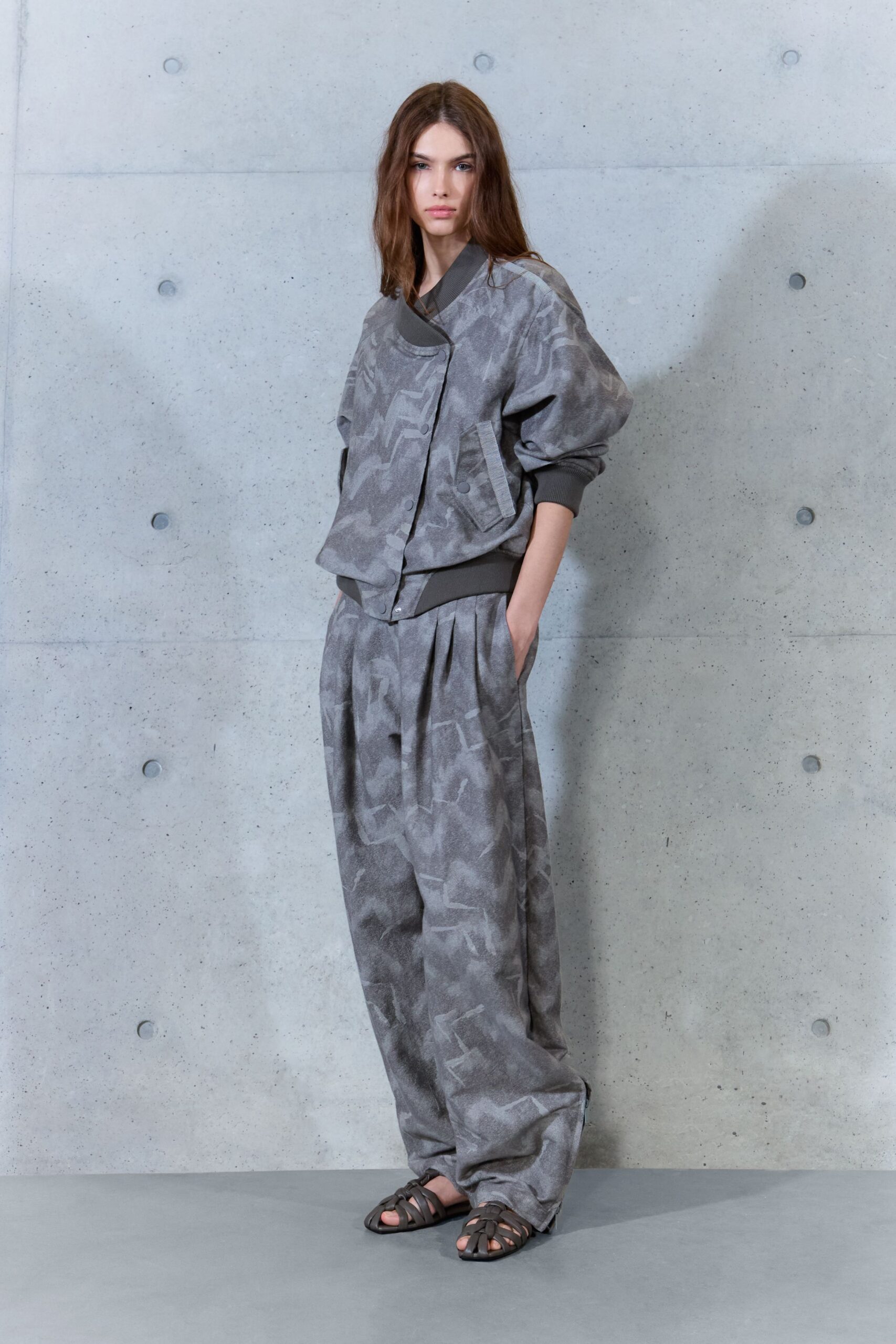 Mottled grey bomber with ribbed collar and snap-flap patch pockets. Matching loose trousers, double pleats falling into a long pooled hem. Dark caged leather sandals. Walking the quiet concrete corridors before the building opens.