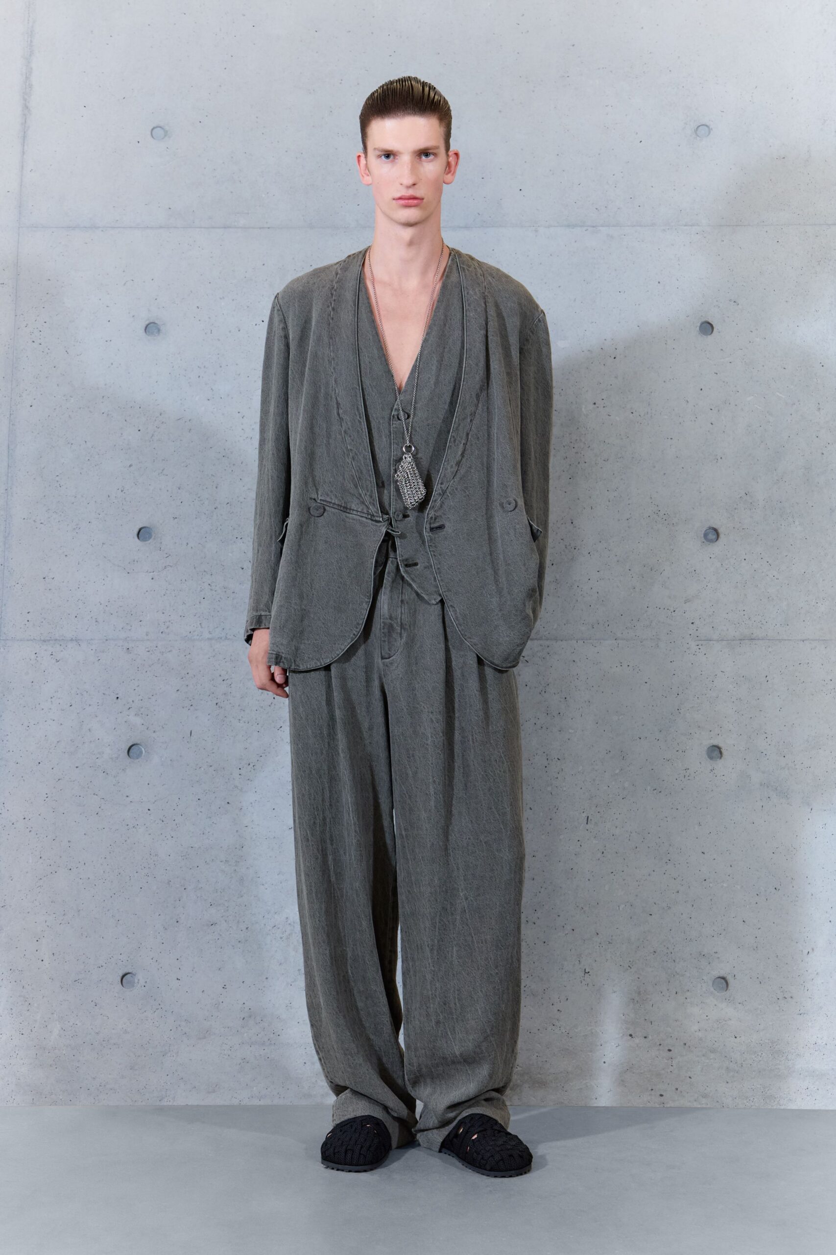 Washed charcoal jacket with curved edges and a low single tie. Deep V vest in the same faded cloth, one button catching low. Small silver mesh pouch hanging from a long chain. Loose pleated trousers pooling over the feet in soft worn twill. Black woven slip-ons. Concrete foyer, early enough that the building still smells damp.