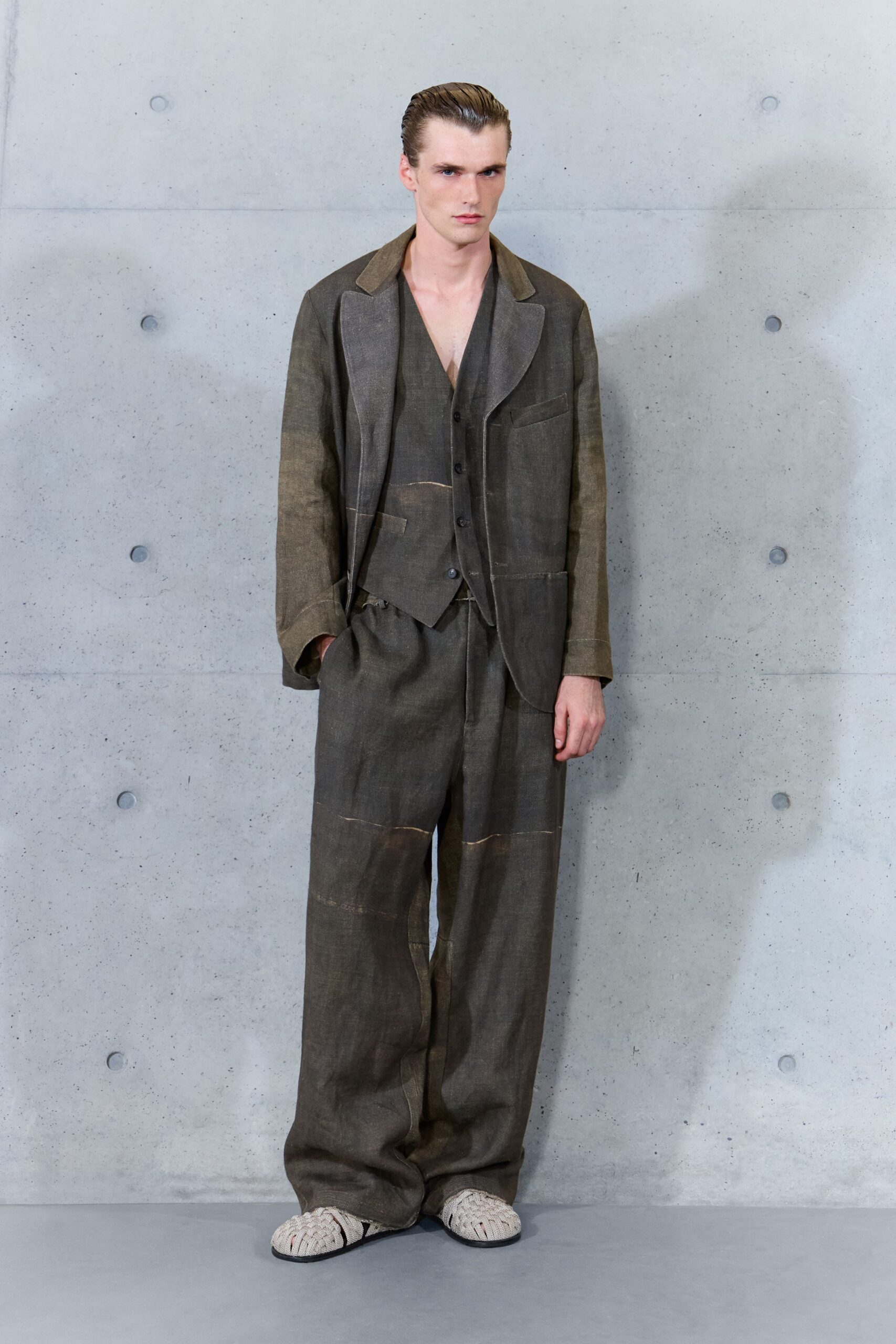 Washed olive jacket with broad raw-edged lapels and oversized patch pockets. Deep V vest in the same slubby cloth, buttoned low. Loose matching trousers with pale seam bands cutting across the legs. Cream woven slippers, black sole peeking at the edge. For crossing a concrete courtyard before the rain actually starts.