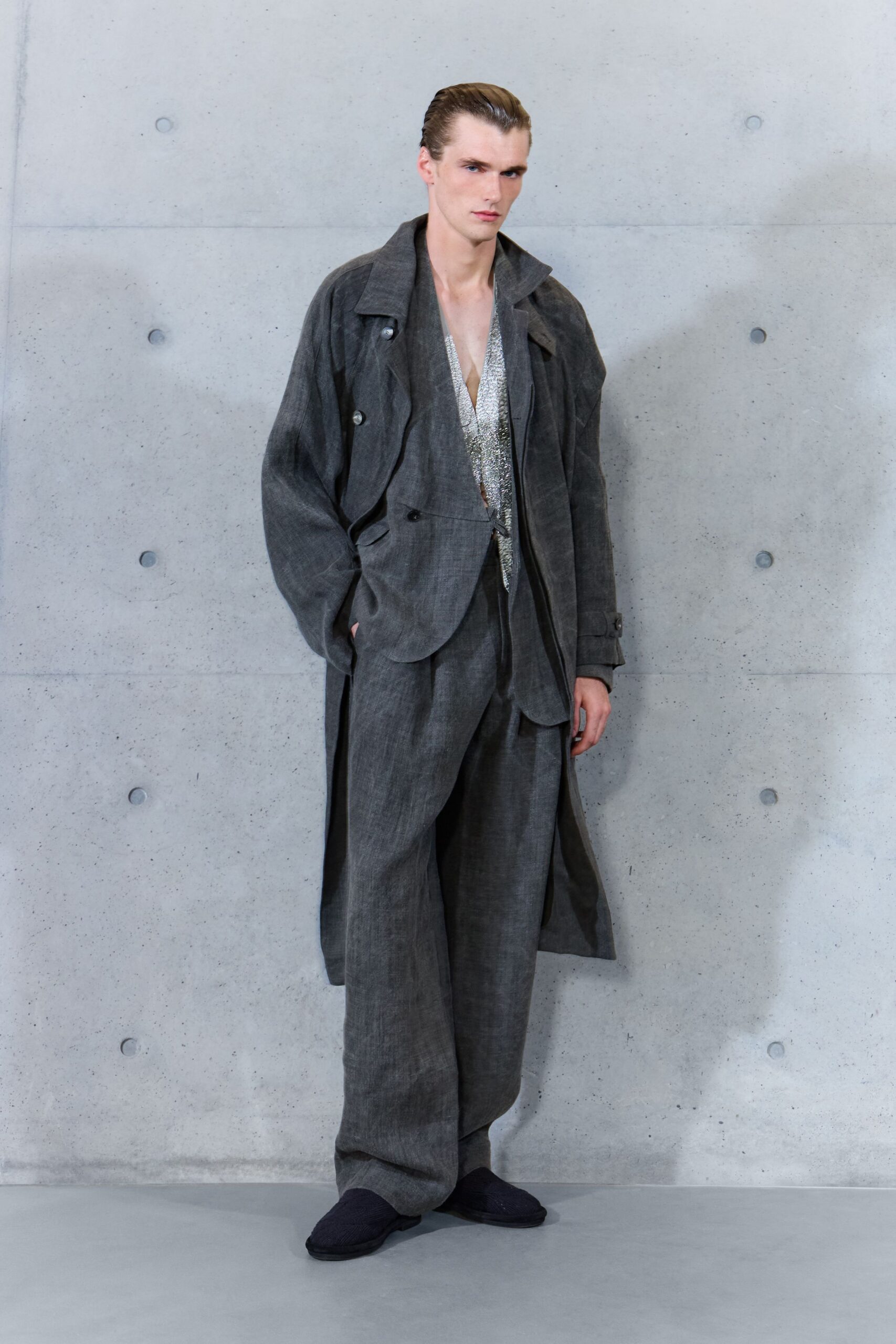 Washed charcoal trench in slubby cloth, one cuff strap hanging loose. Silver chainmail plunge top catching light inside the open coat. Wide charcoal trousers in the same dry cloth, pooling over the foot. Dark knit house slippers. For the quiet elevator ride down after a hotel bar emptied out.