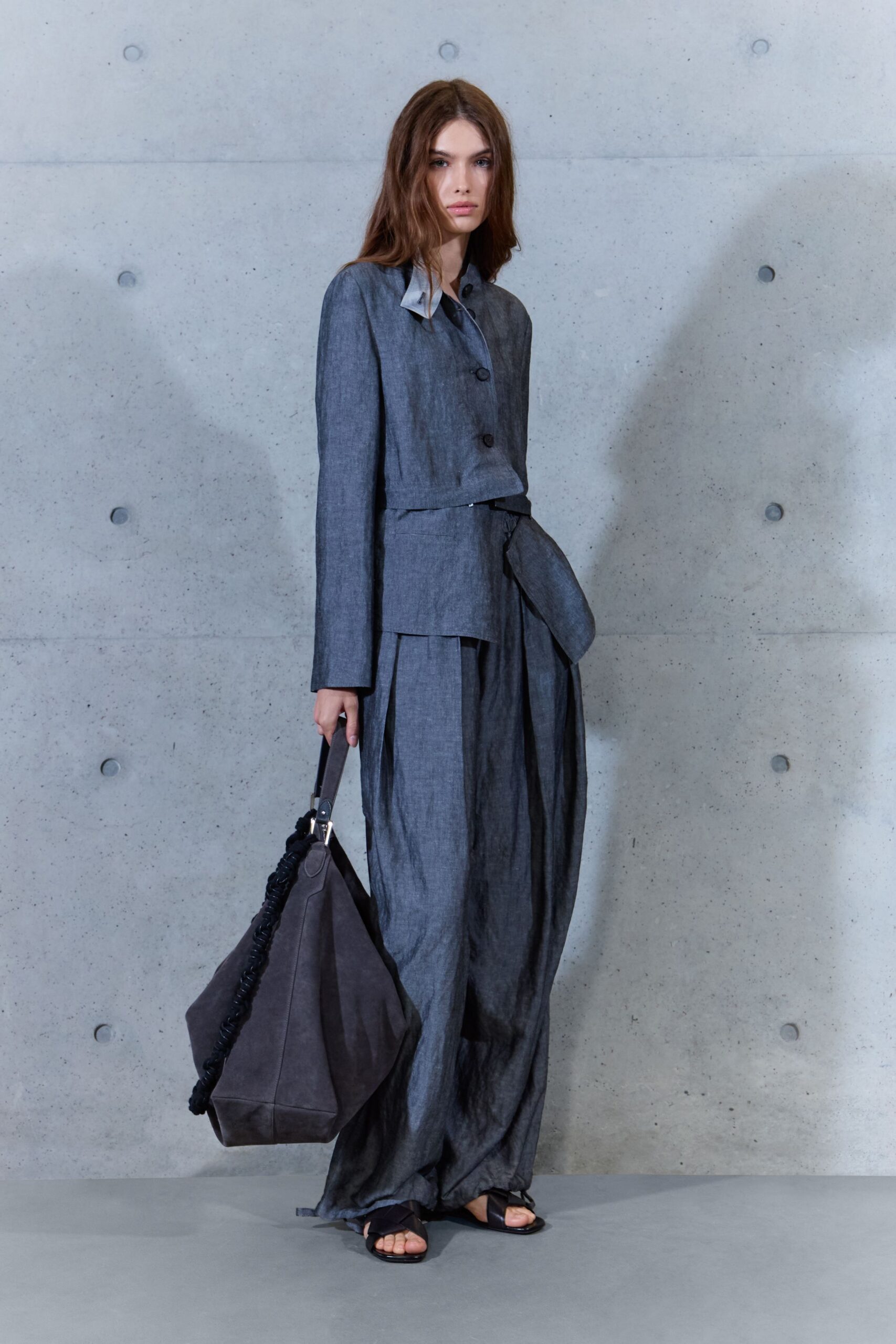Cropped slate chambray jacket with a pale undercollar and two oversized buttons. Matching floor-length wrap skirt, one apron panel falling loose at the hip. Charcoal suede tote with a ruched black edge. Black leather thong sandals. For crossing a silent concrete lobby after the rain has stopped.
