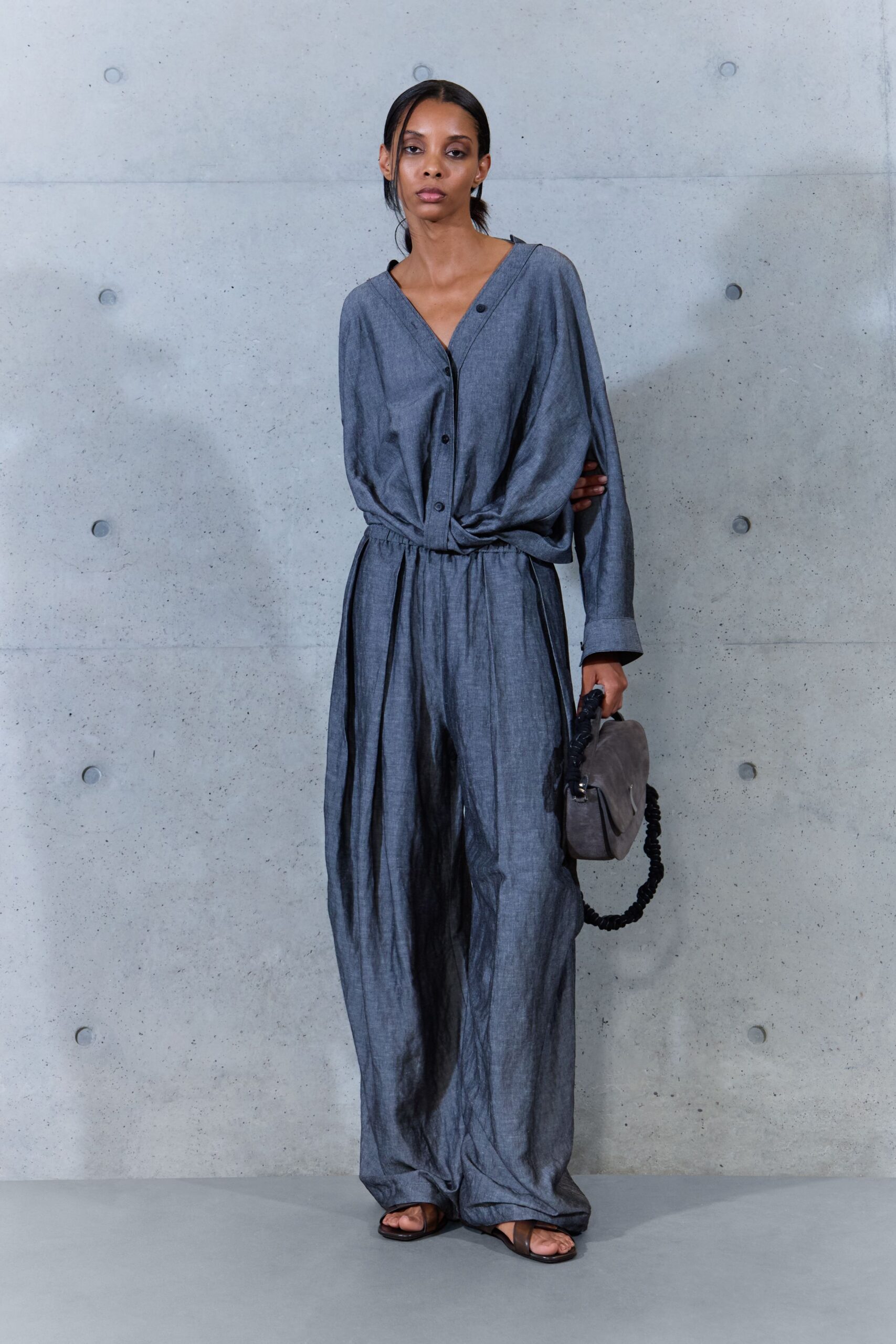 Slate chambray jumpsuit with a deep V, dropped shoulder, and gathered waist. Taupe suede shoulder bag with a ruched black strap. Flat brown leather sandals, barely visible under the pooling hem. Whatever you wear home after the ferry deck wind has gone warm.