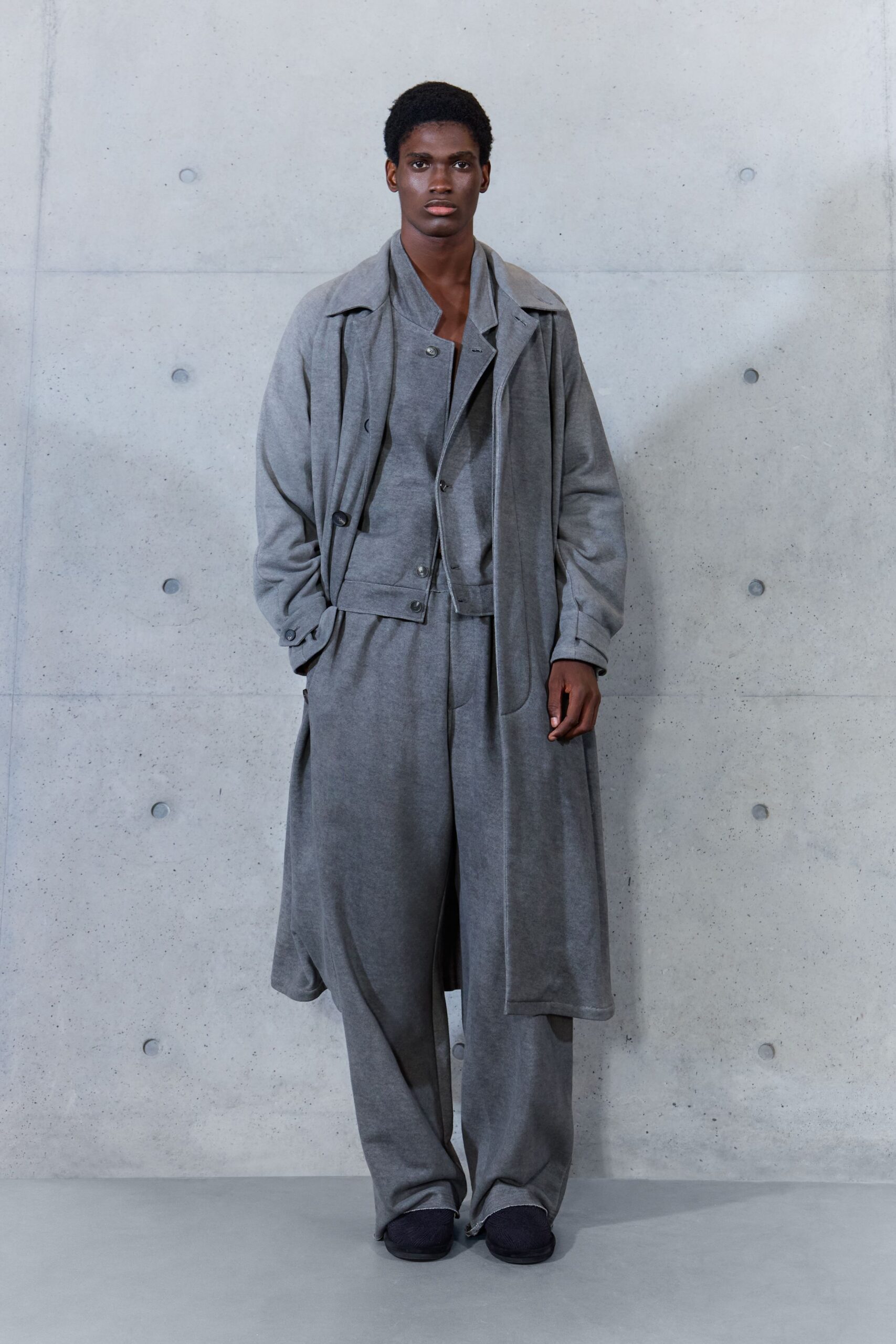 Long heather-grey jersey coat with a limp self belt hanging loose. Matching knit jacket, one snap left open below the deep V. Wide grey trousers in the same soft marl, pooling at the hem. Dark slip-ons, rounded and almost swallowed by the break. Waiting out a drizzle under the library overhang after closing.
