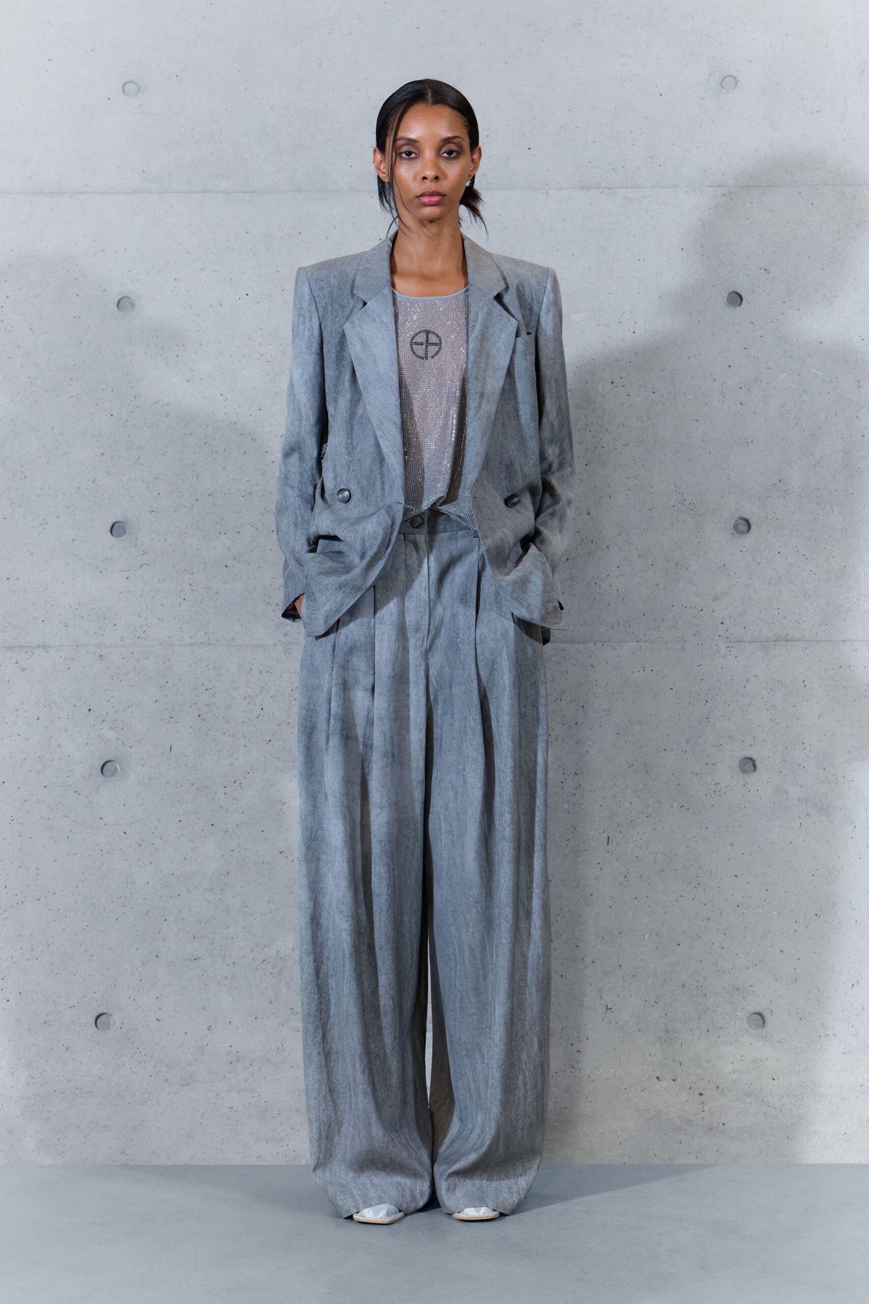 Washed grey blazer with peaked lapels and low doubled buttons. Silver mesh sleeveless top, a black circle-cross emblem at the chest. Matching wide trousers with deep front pleats breaking over the shoes. Pointed silver shoes peeking out under the pooled hem. Early office on a Sunday, concrete still holding the night's cold.