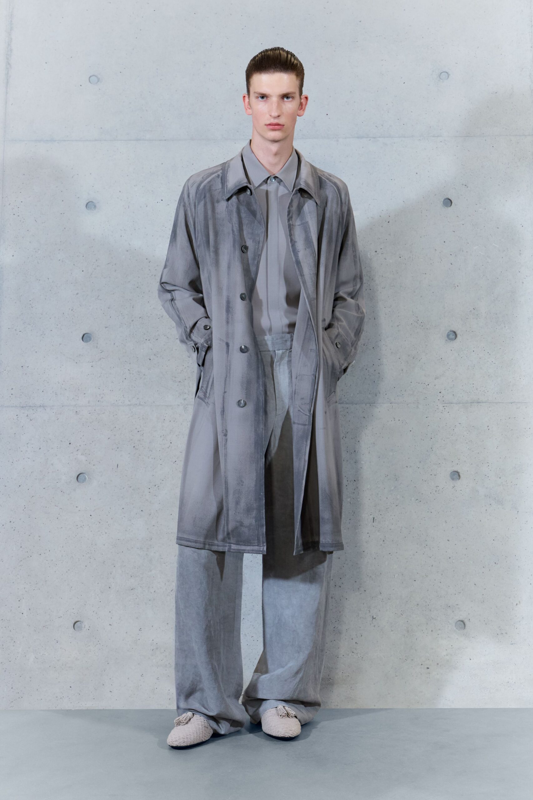 Sheer dove-grey coat with watery brushed shading and dark horn buttons. Matching grey shirt, placket hidden beneath a narrow center strip. Loose pale grey trousers with a soft crushed nap. Cream woven slippers with tassels at the vamp. Waiting out a spring downpour under the museum loading dock.