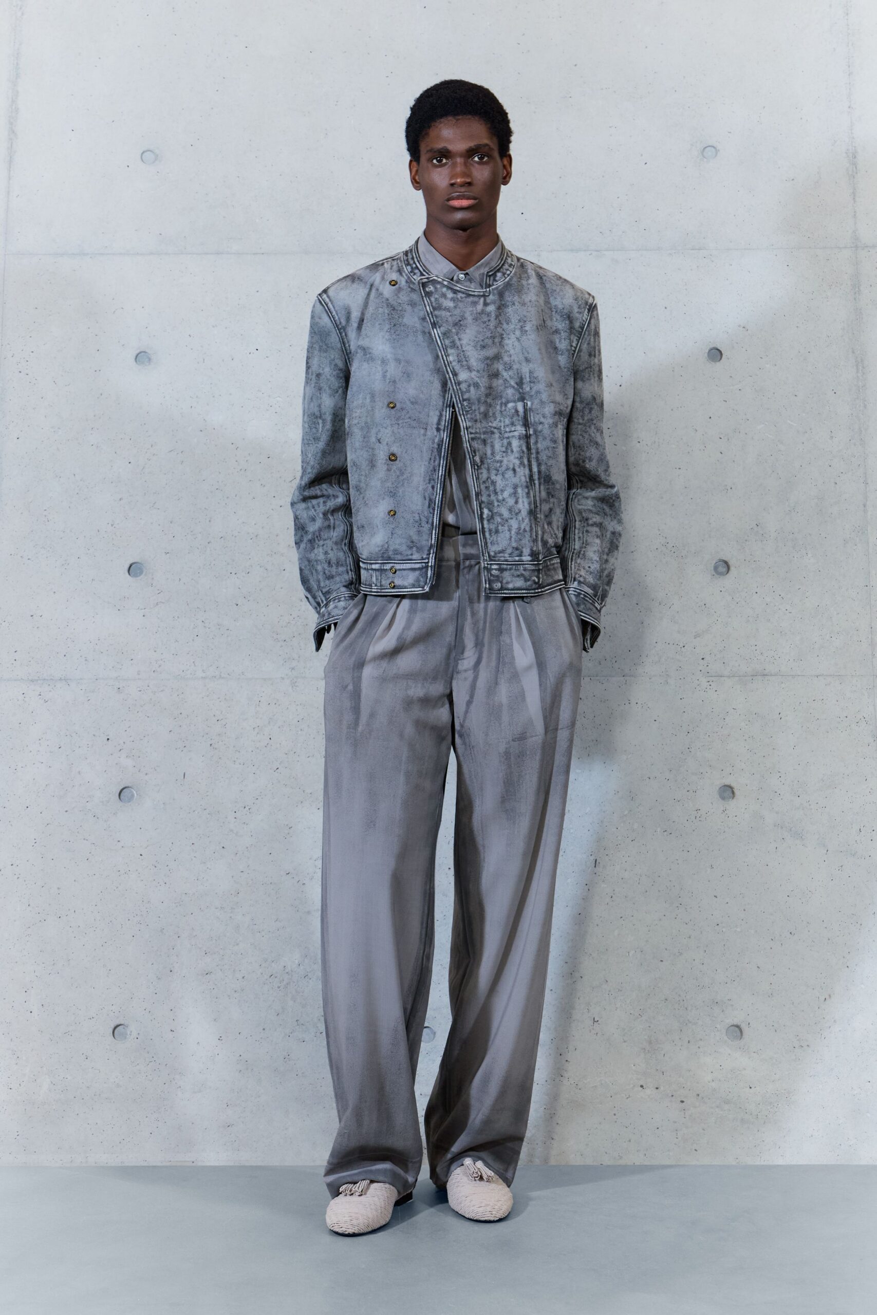 Mottled grey jacket with an off-center placket and brass snaps. Pale grey shirt buttoned tight at the small stand collar. Fluid dove-grey trousers, sharply creased and pooling over the shoe. Cream woven slippers with a knotted tassel at each toe. Early train to the city, concrete still holding last night's cold.